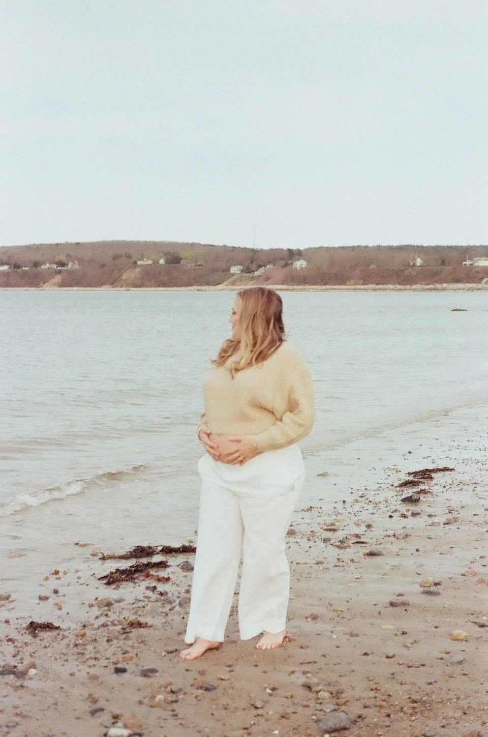 A woman with long hair standing on a beach, facing the water, with her hands resting on her belly, wearing a beige sweater and white pants.