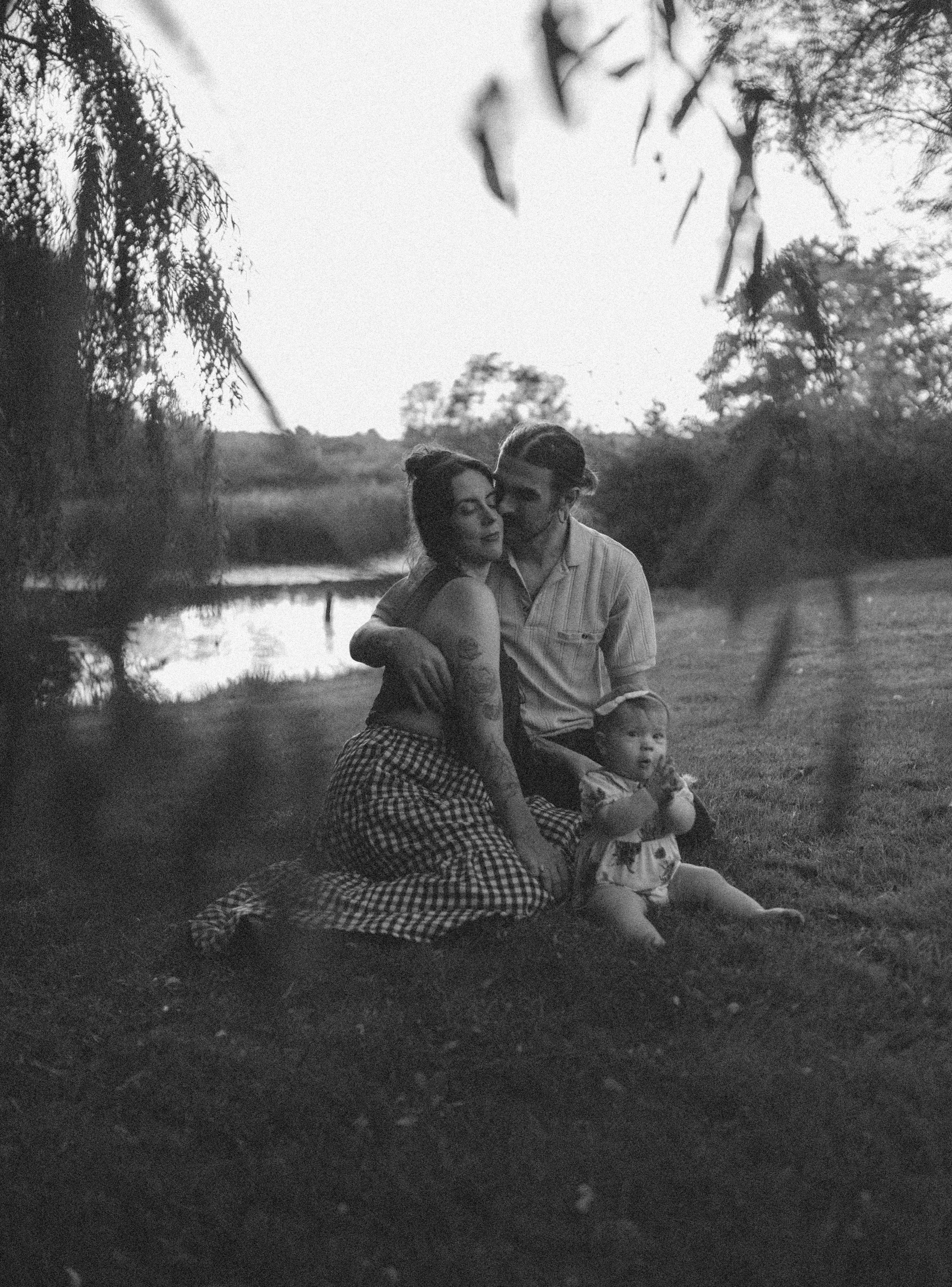 A black and white photo of a family at a lakeside during sunset. A woman with tattoos and a checkered skirt is sitting on the grass, leaning into a man who is kissing her forehead. A young child is sitting in front of them, holding a toy.