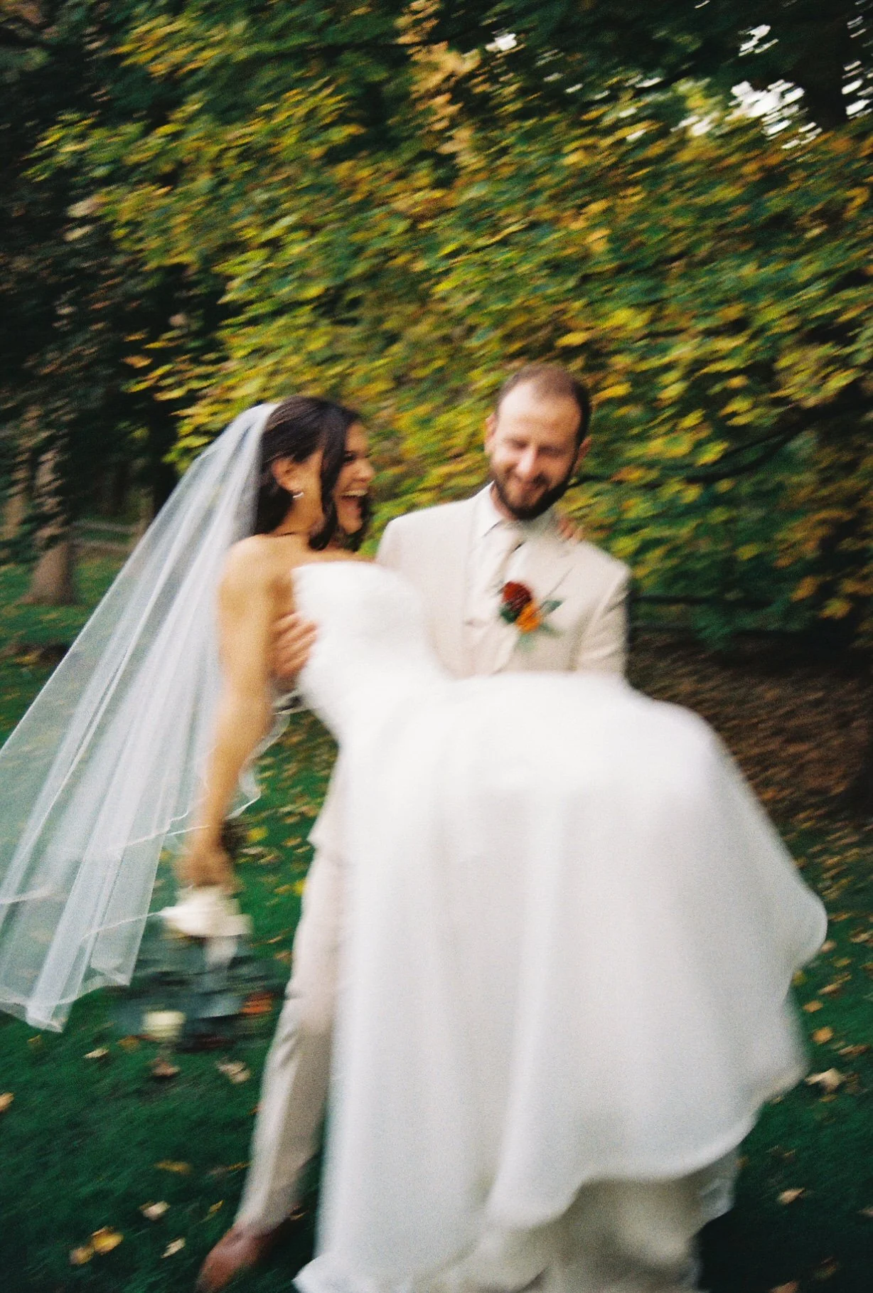 A bride and groom smiling and laughing outdoors, with the groom holding the bride in a white wedding dress, surrounded by autumn foliage.