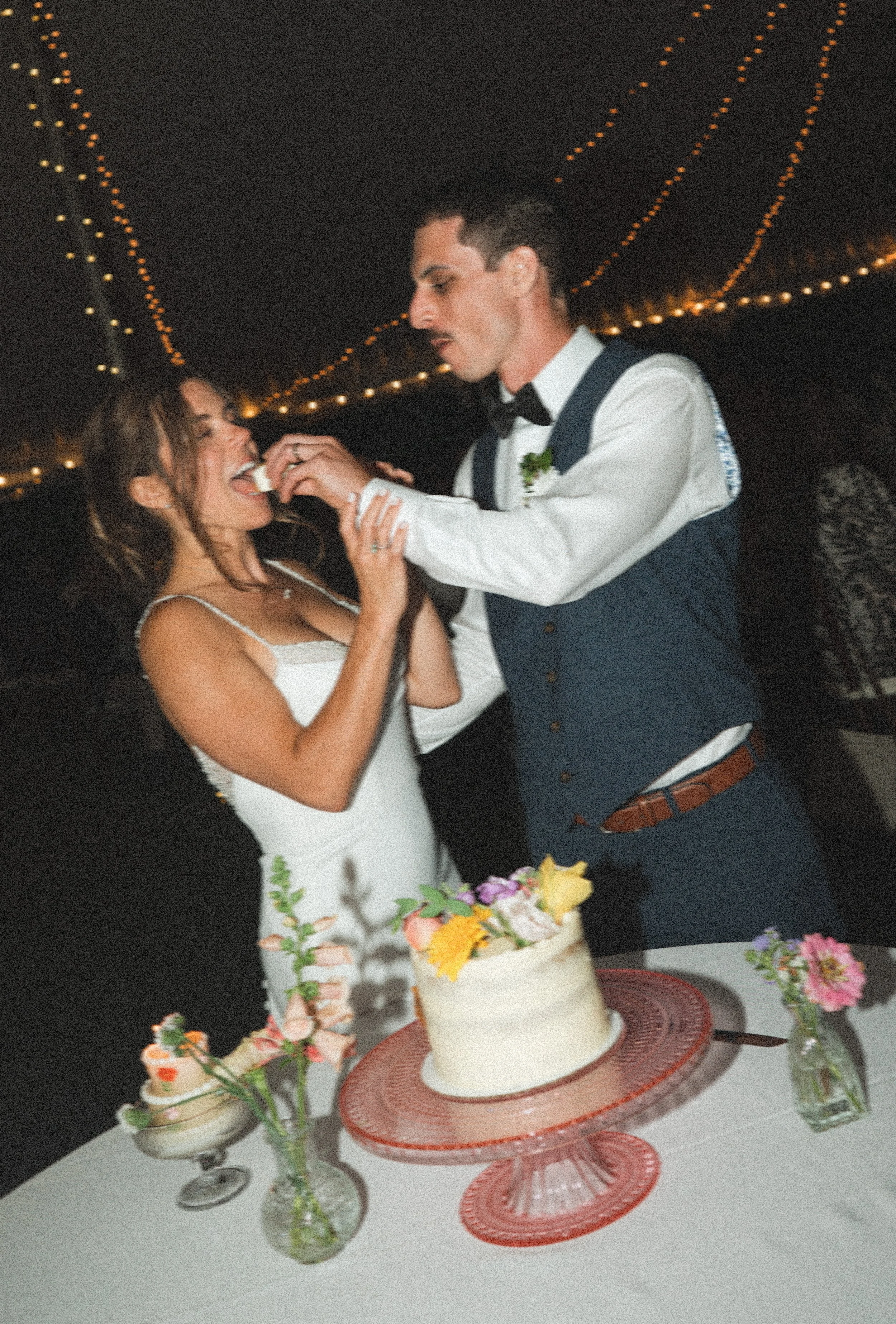 A newlywed wedding reception with a bride and groom feeding each other cake under string lights.