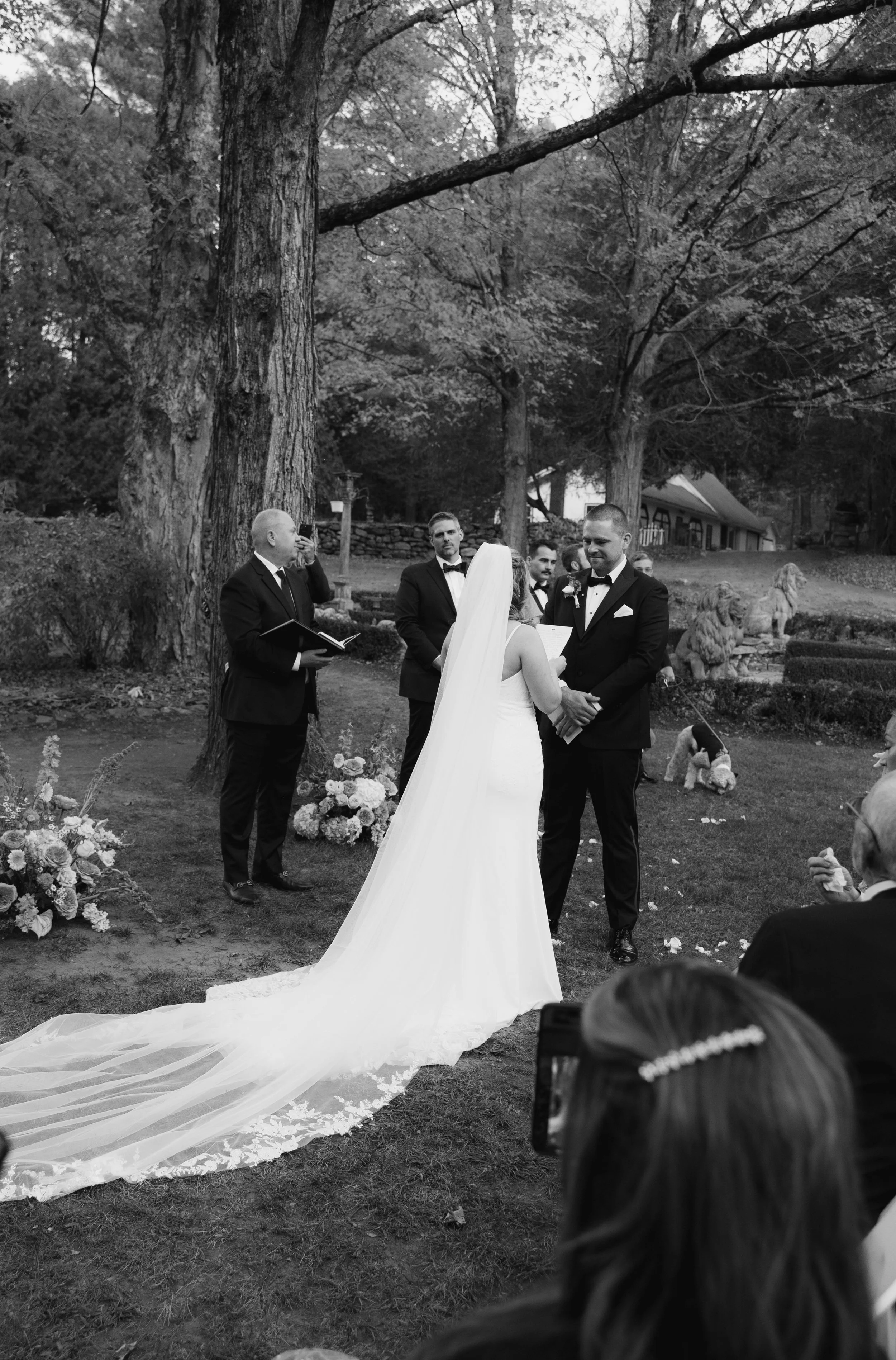 A wedding ceremony outdoors with the bride and groom holding hands, surrounded by officiants and guests, with large trees and a house in the background.