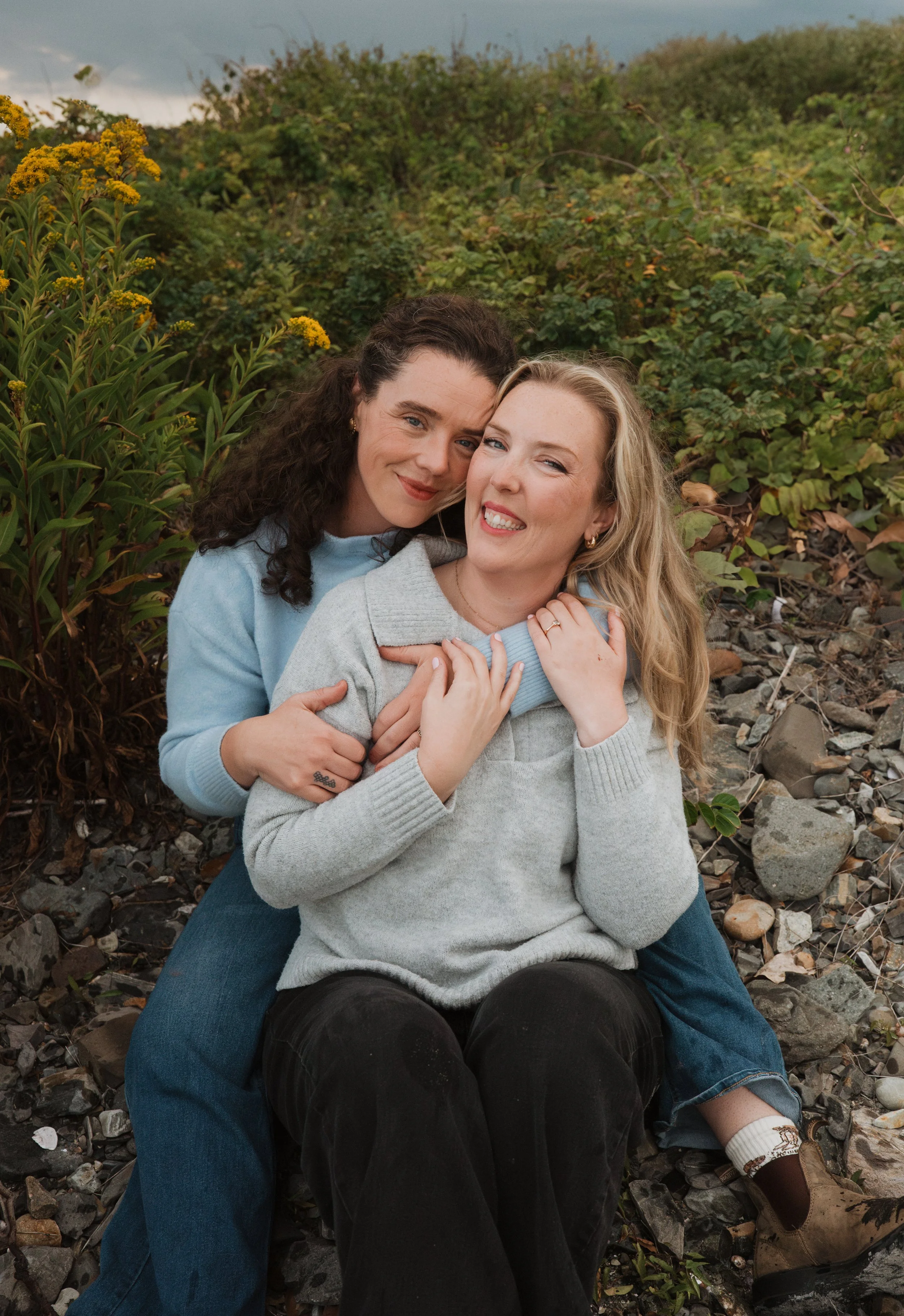Two women embracing outdoors on a rocky ground, surrounded by greenery and yellow flowering plants, with cloudy sky in background.