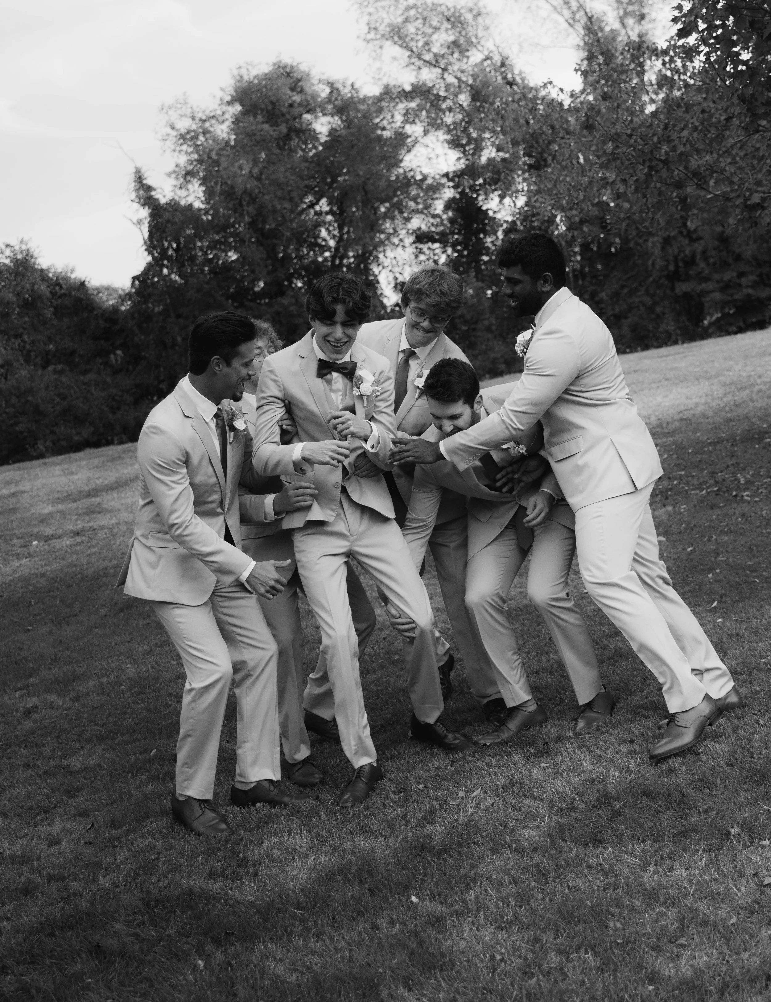 Group of six men in suits, laughing and playing on a grassy field during daytime.