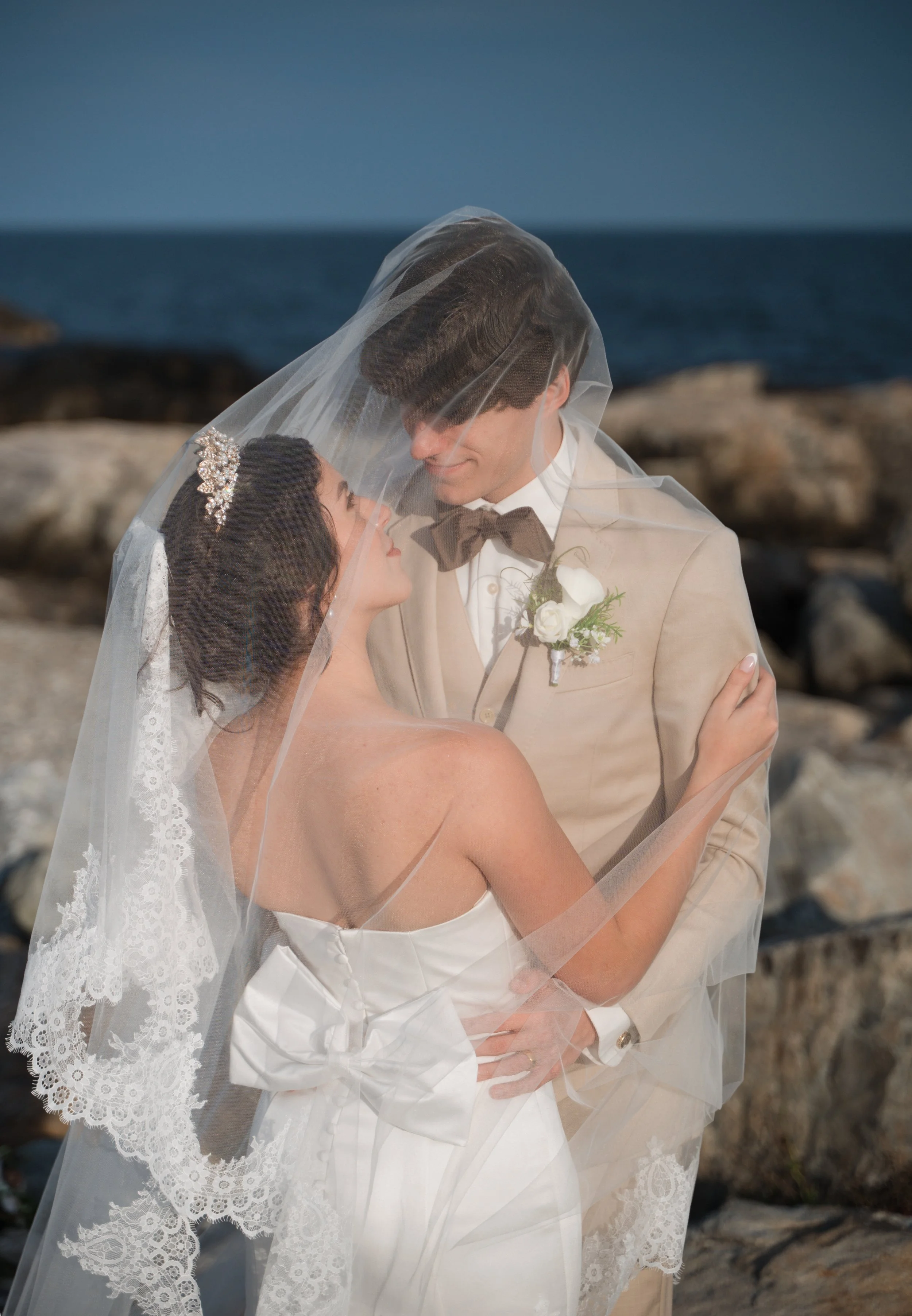 A bride and groom dressed in wedding attire, embracing on a rocky shore near the ocean, with the bride's veil covering both of them.