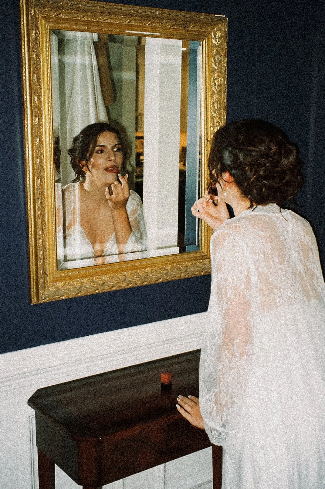 A woman in a white lace robe applying makeup while looking at her reflection in an ornate gold-framed mirror.