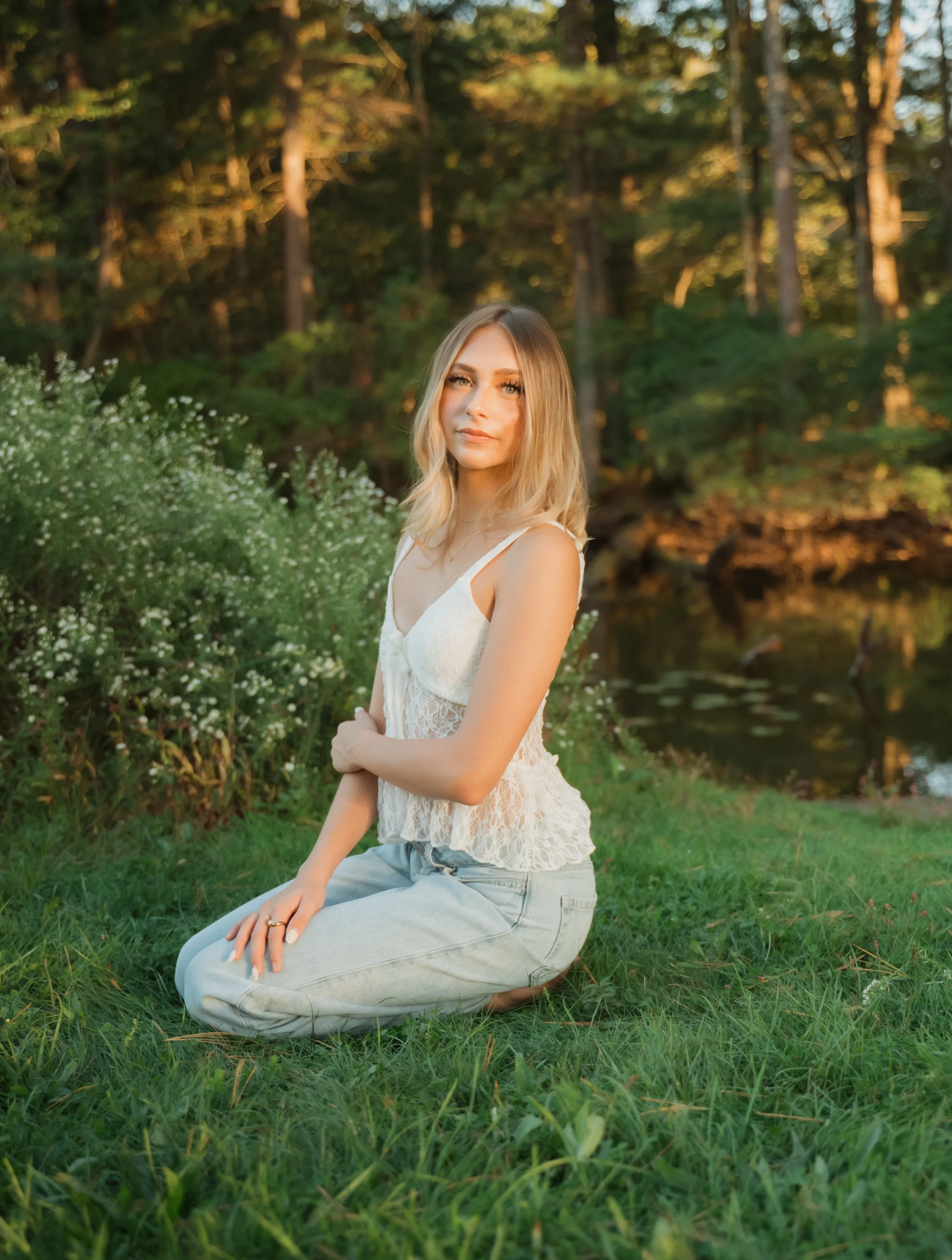 A young woman with blonde hair sits on green grass near a pond, with trees in the background, during sunset.