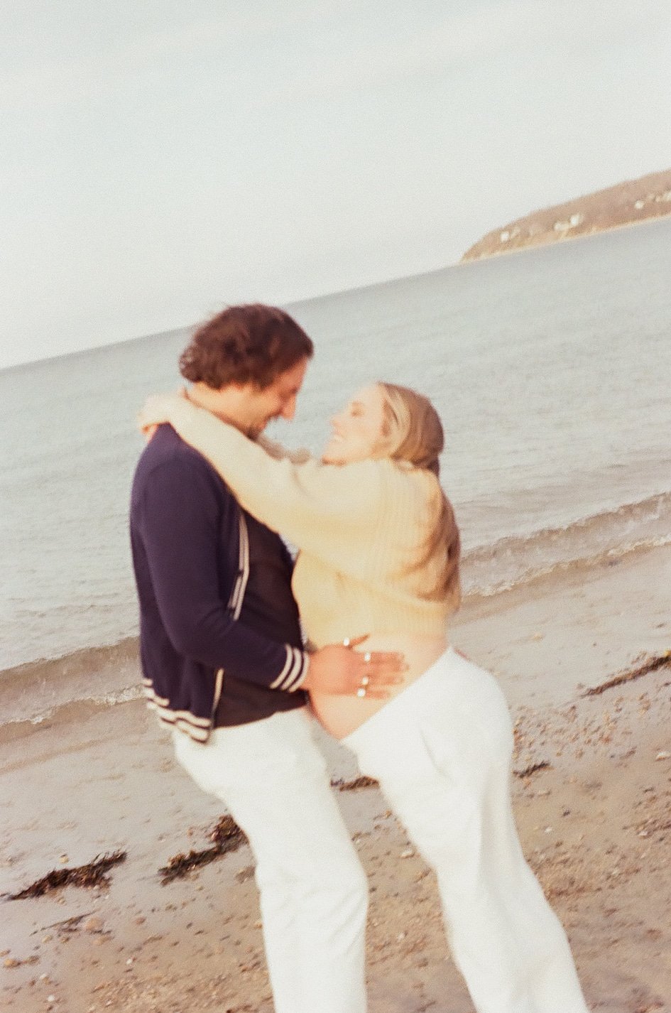 A couple enjoying a romantic moment on the beach, embracing each other and smiling, with the ocean and a distant landmass in the background.