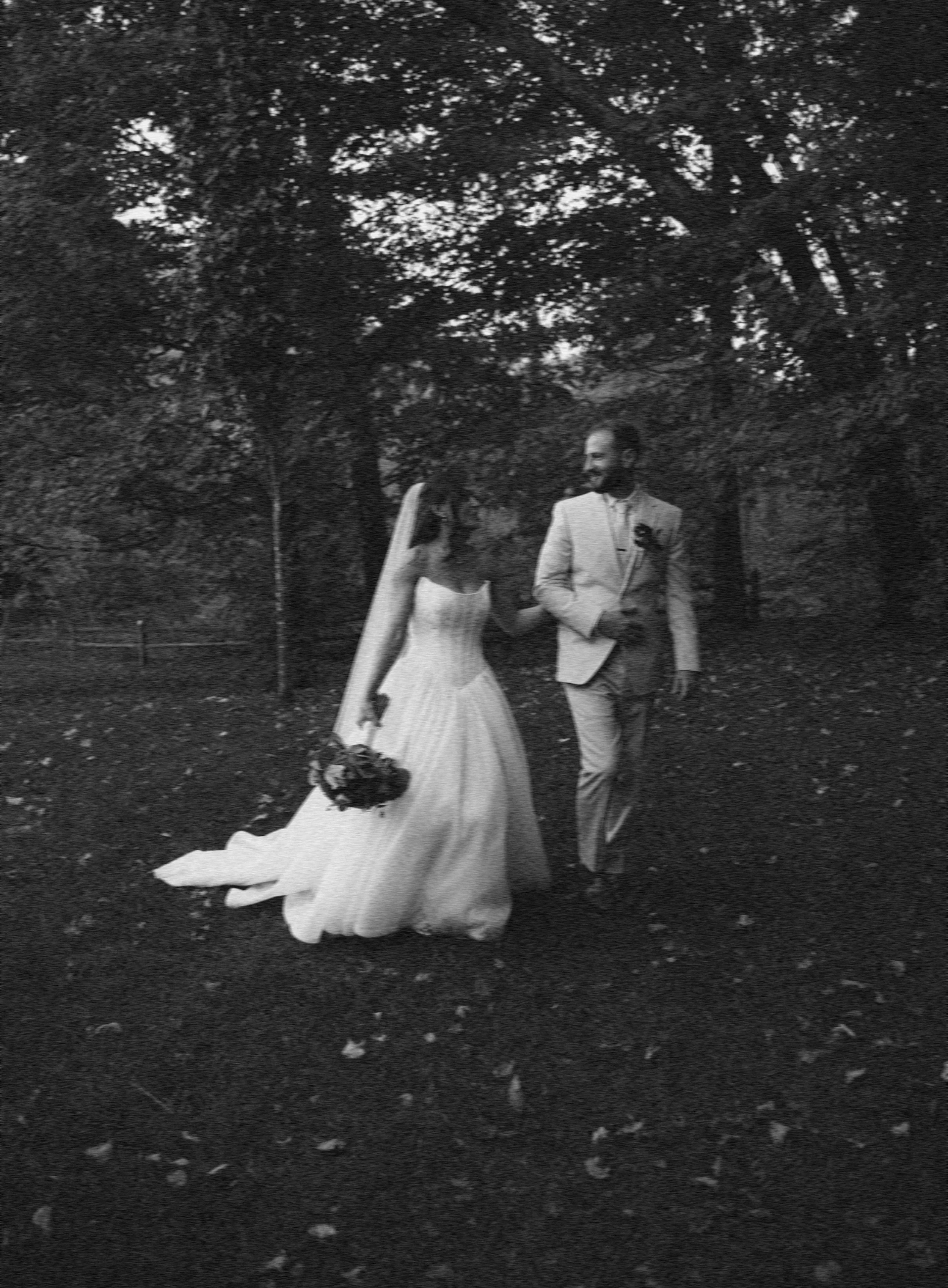A bride and groom walking hand in hand outdoors beneath trees, dressed in wedding attire.
