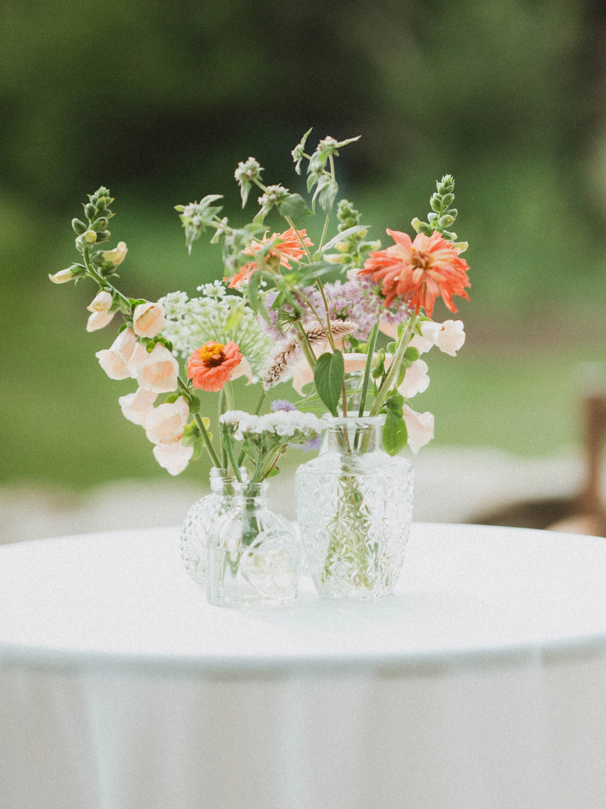 A bouquet of various colorful flowers in two textured glass vases on a white table, with a green blurred background.