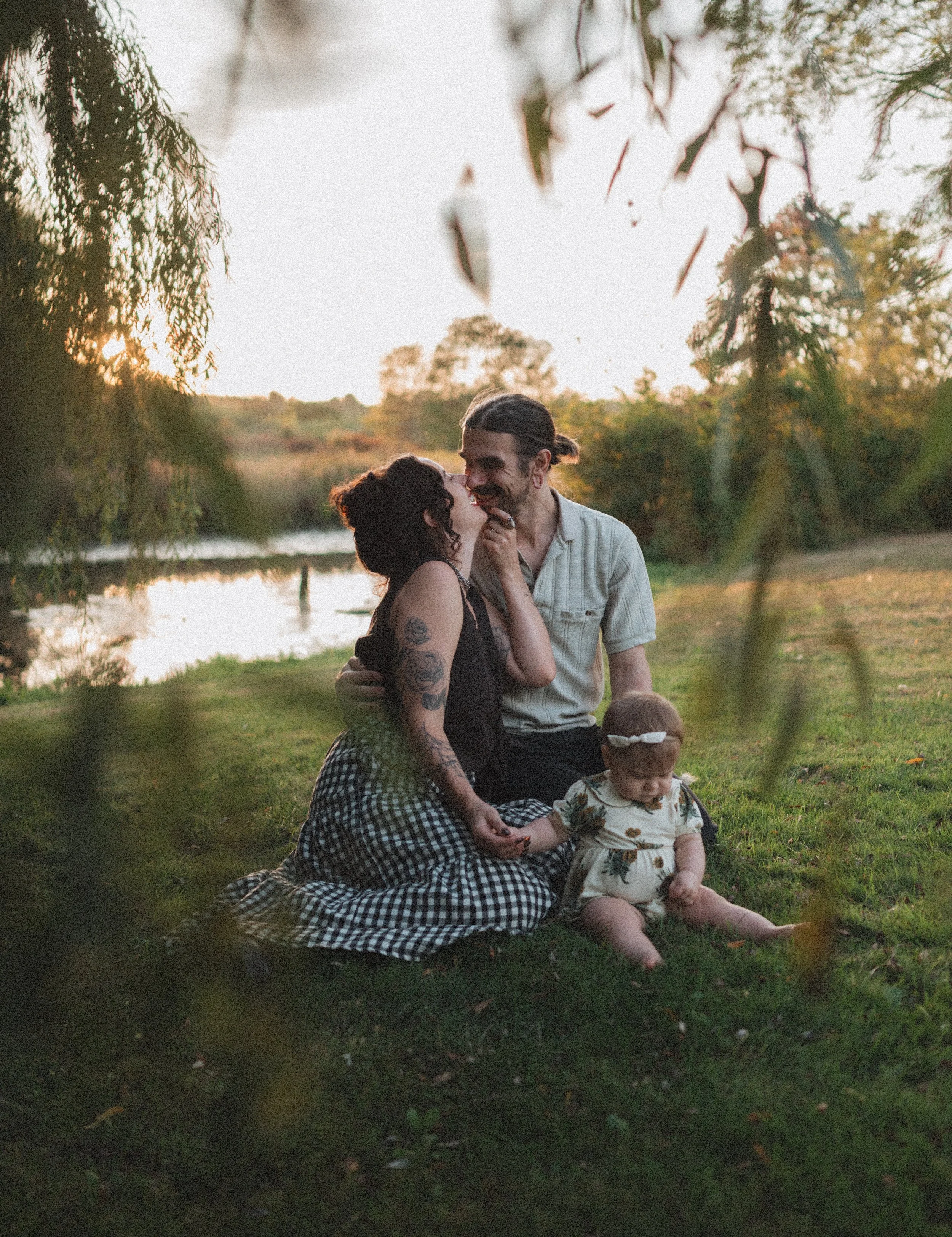 A family of three sitting on grass near a river at sunset, with trees in the background, the woman and man smiling and holding hands, and a young girl sitting on the ground, holding the woman's hand.