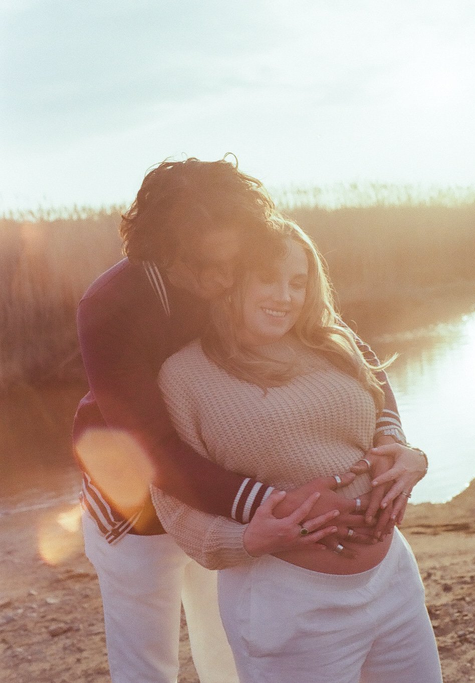 A couple standing on a beach near water, embracing each other and smiling at sunset.