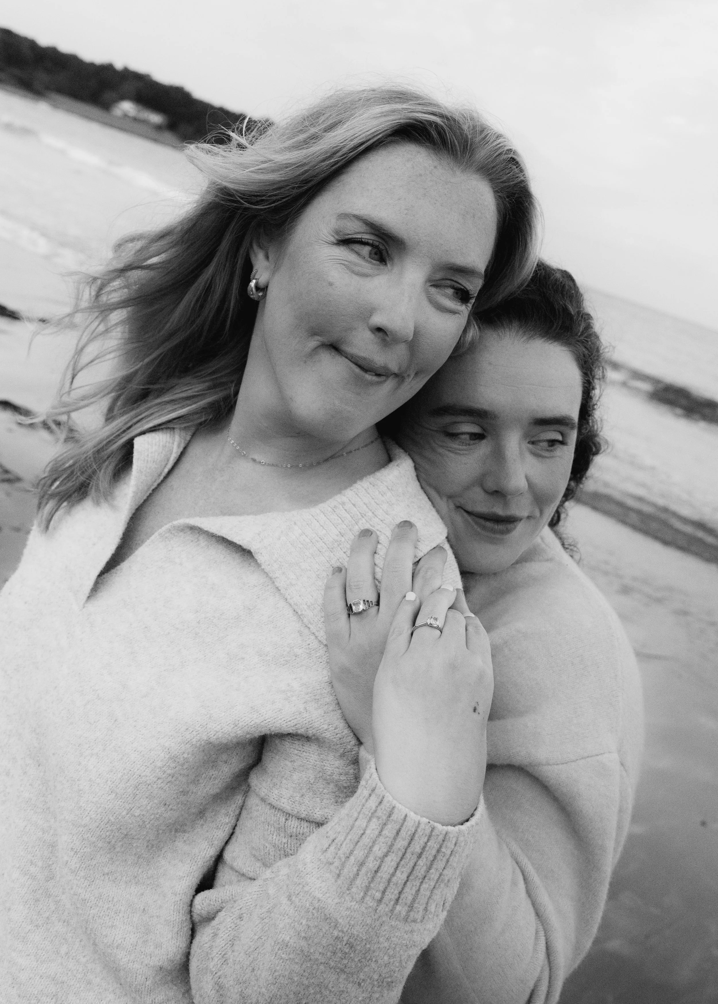 Two women hugging on the beach, with the ocean and sky in the background, one woman with light hair and the other with dark curly hair, both wearing sweaters, in black and white.