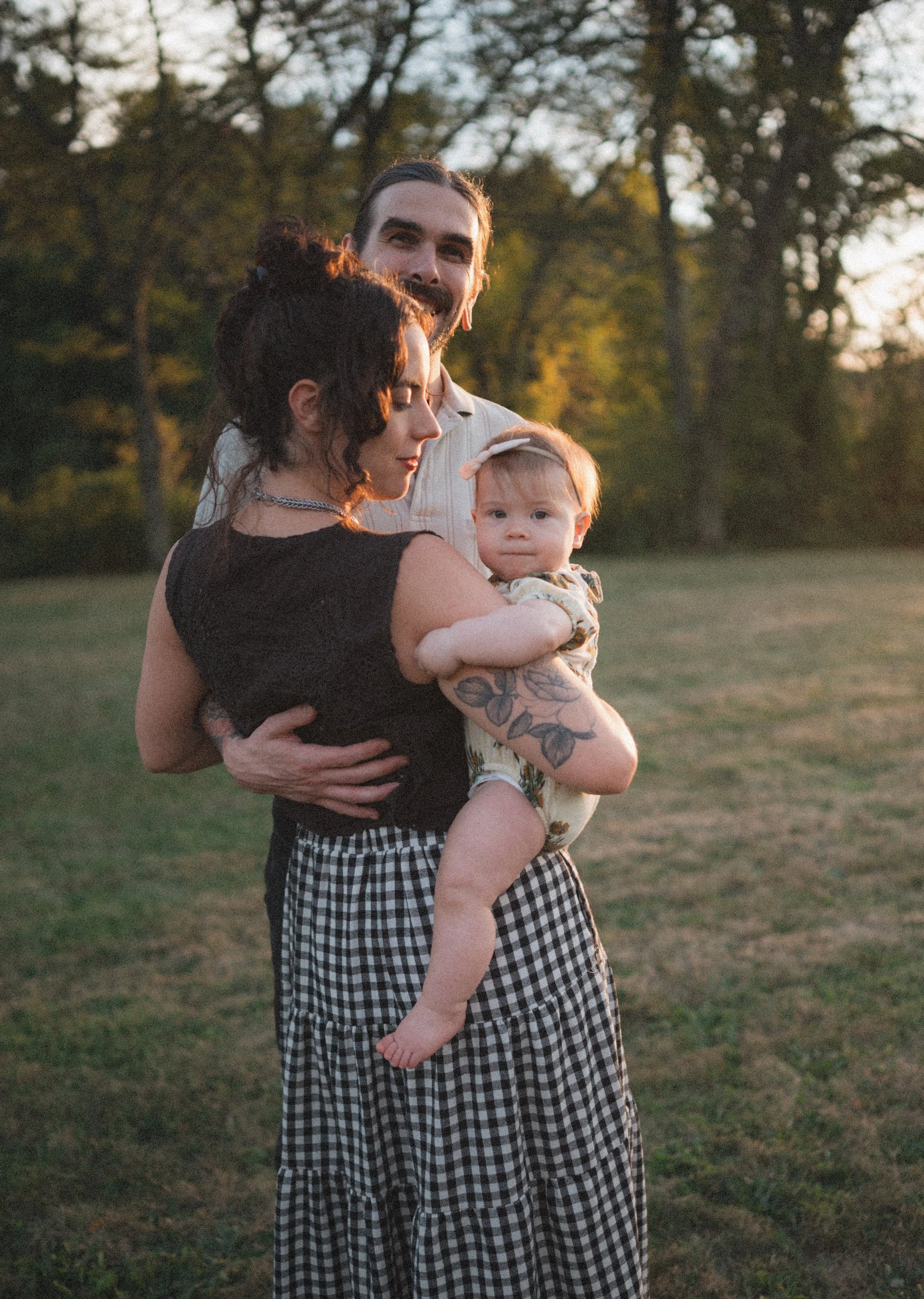 A family of three standing outdoors during golden hour, with trees in the background. The man is smiling, holding the woman and a baby girl. The woman has dark curly hair, is wearing a black sleeveless top and a checkered skirt. The baby girl has light hair, a bow on her head, and is wearing a patterned romper.