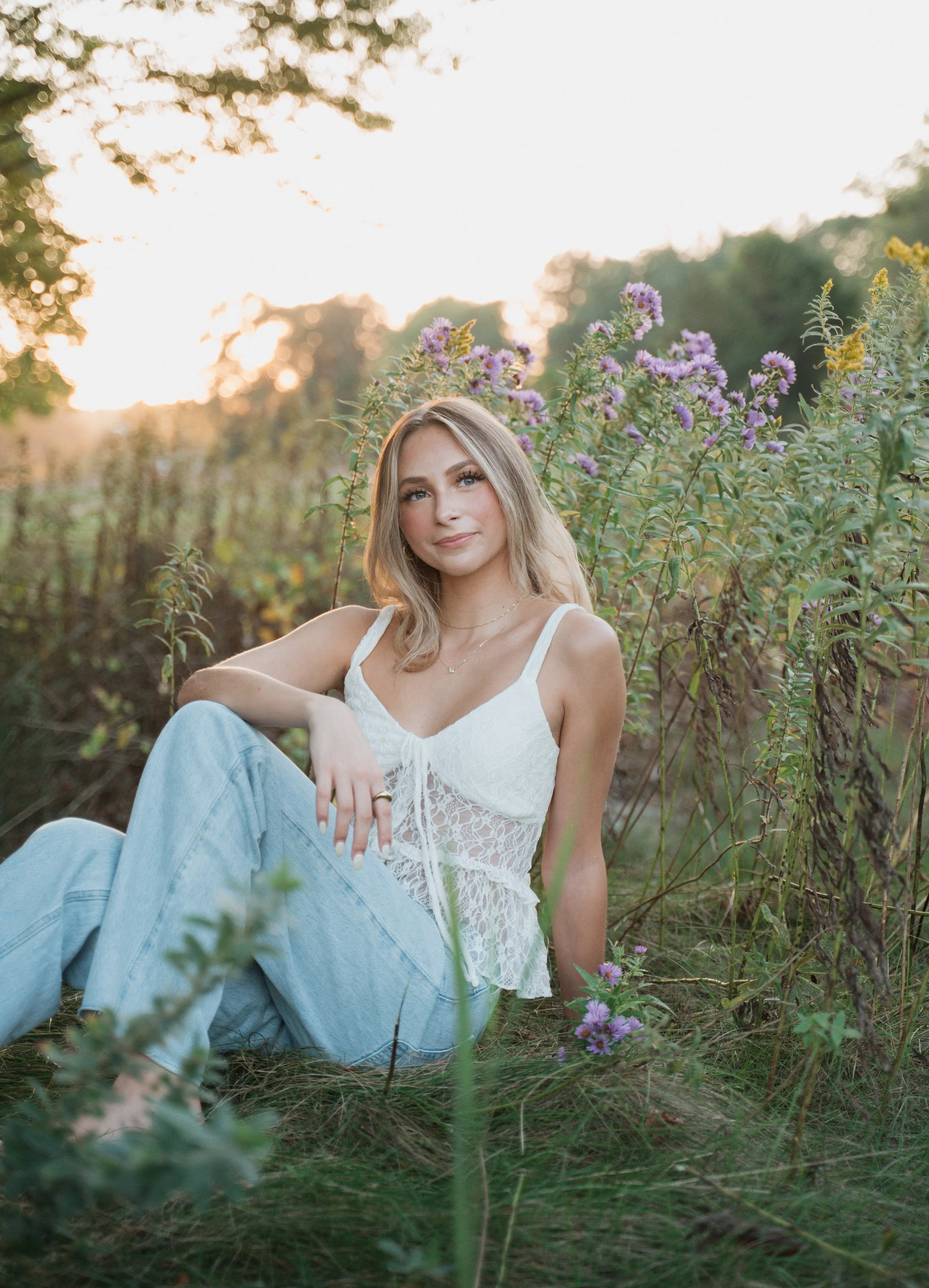A young woman with blonde hair sitting on the grass in a field of purple flowers during sunset. She is wearing a white lace top and light blue jeans, looking at the camera with a slight smile.