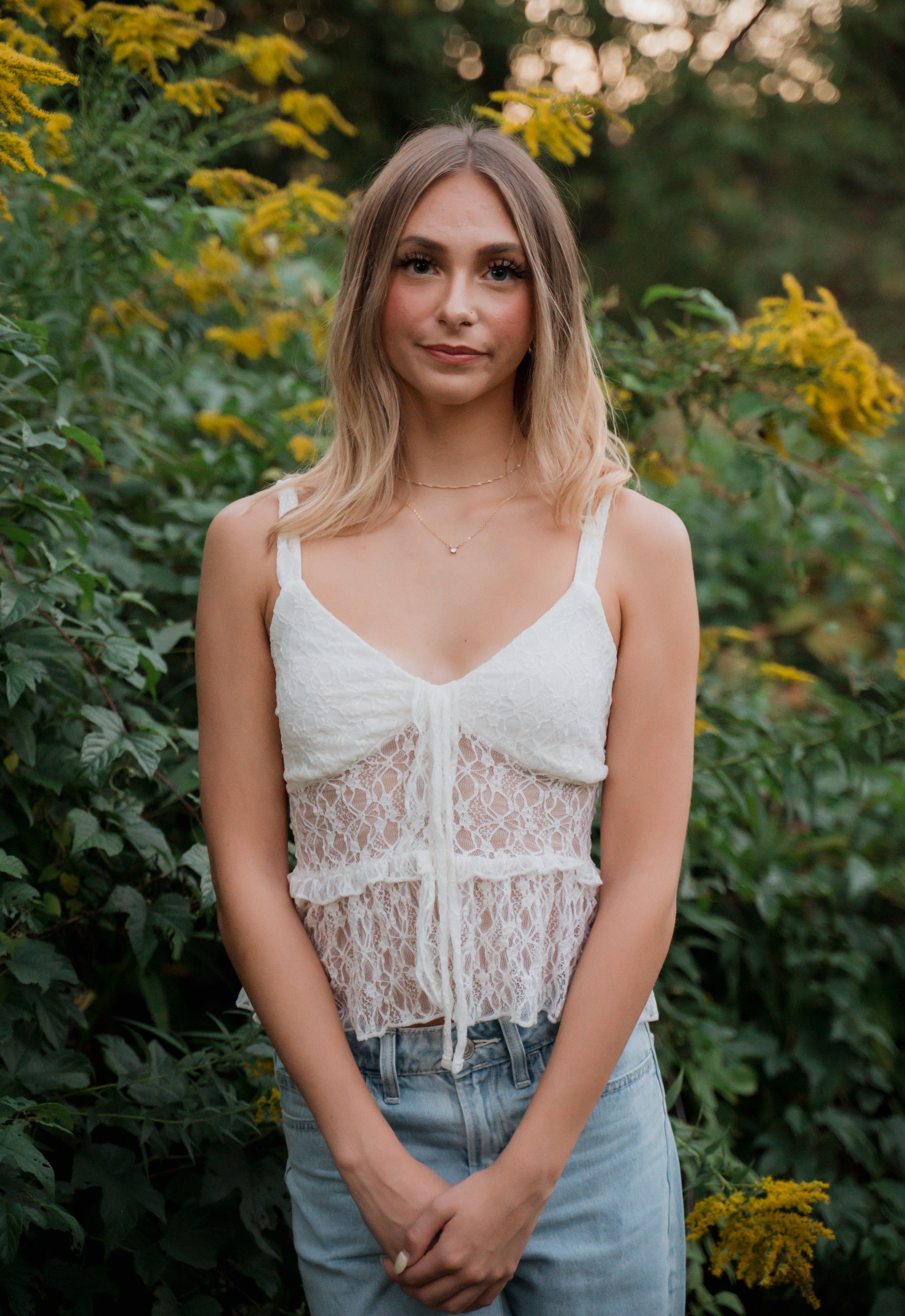 A young woman with shoulder-length blonde hair standing outdoors in front of green foliage with yellow flowers, wearing a white lace tank top and light blue jeans.