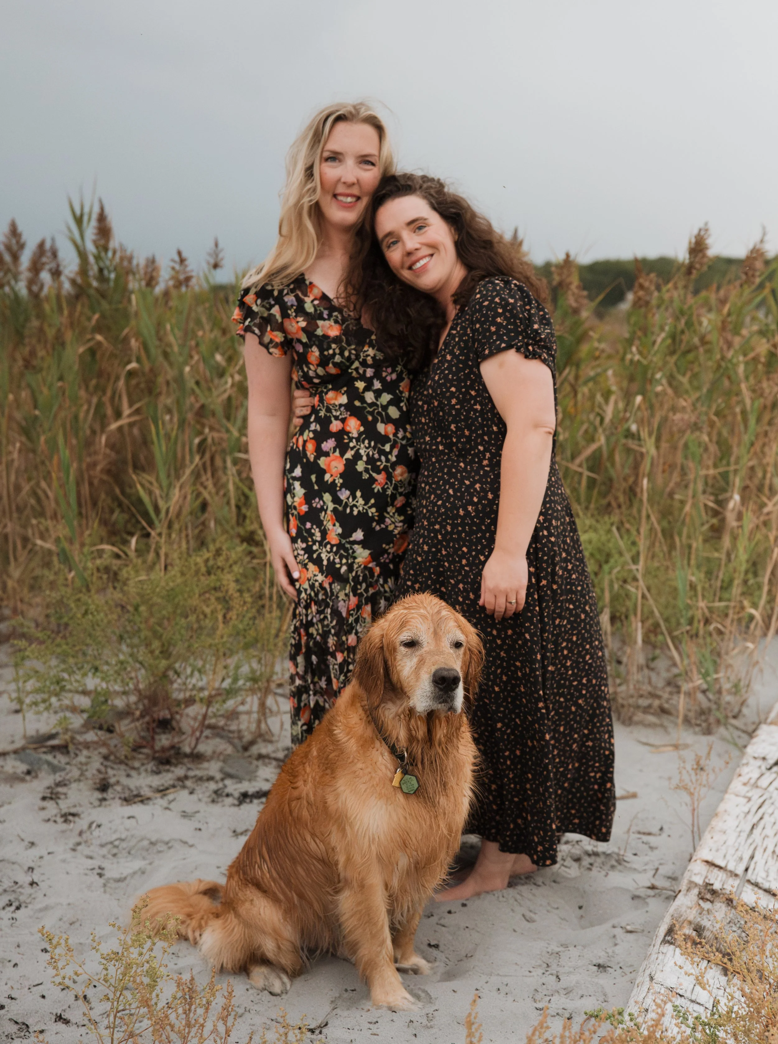 Two women and a dog at the beach, with tall grass in the background. The women are smiling, and the dog is sitting on the sand in front of them.