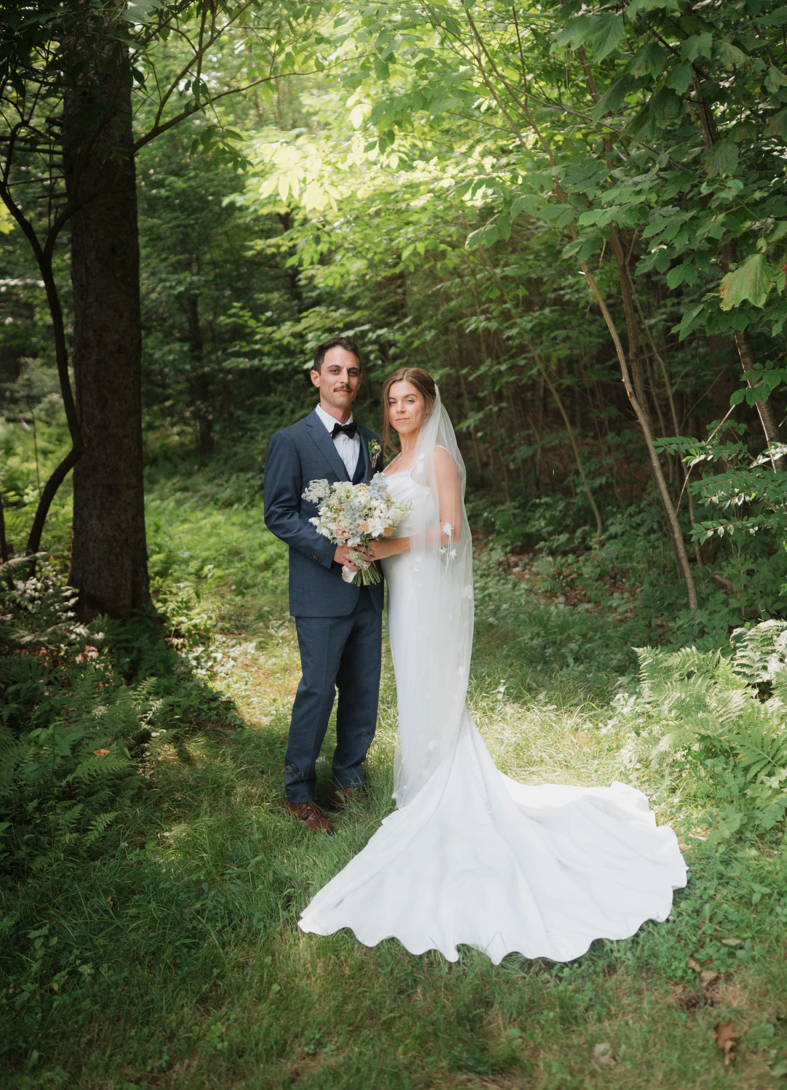 A bride and groom standing together in a forest, the groom in a blue suit with a bow tie and the bride in a white wedding dress with a long train, holding a bouquet of flowers.