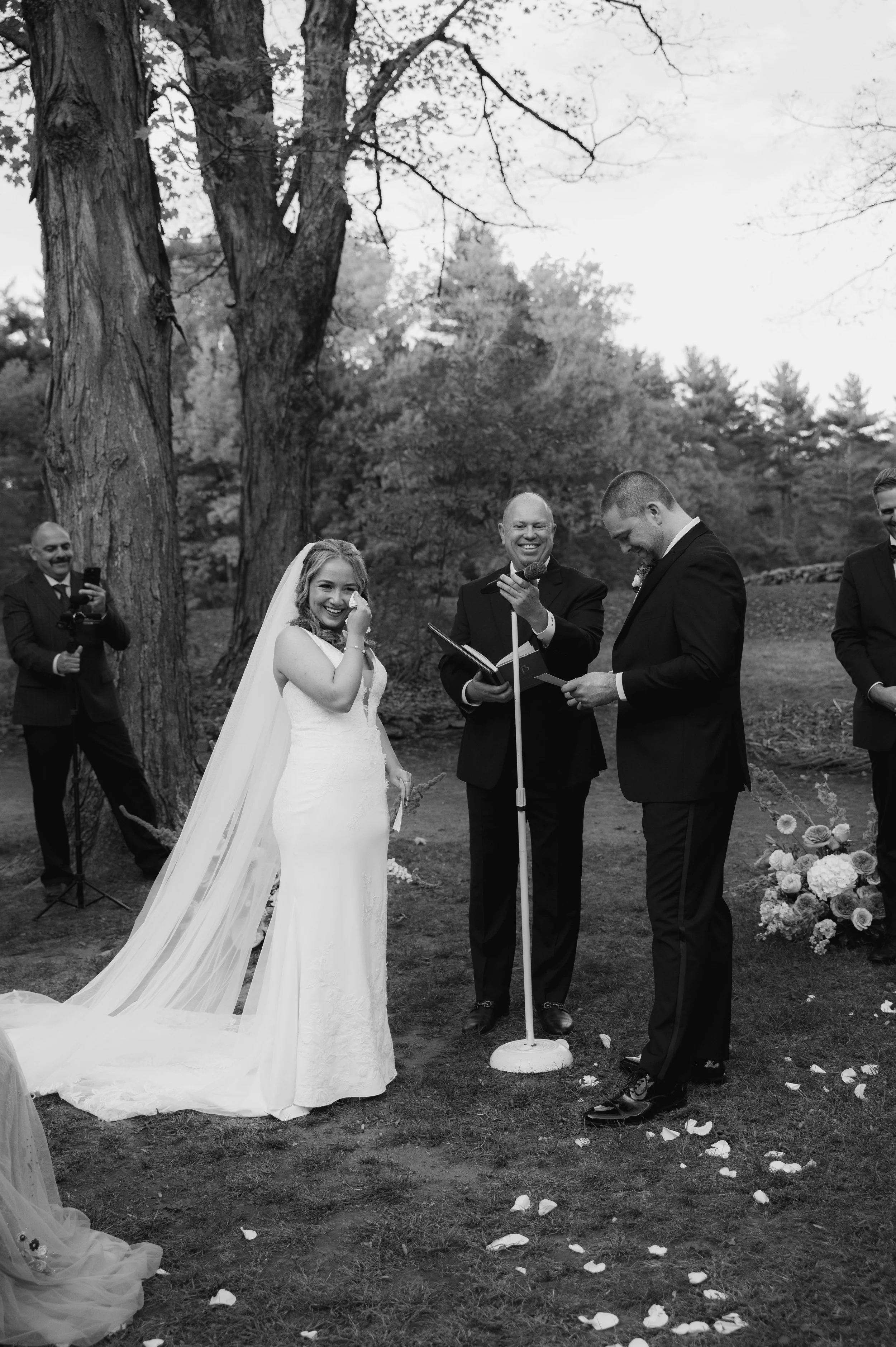 Black and white photo of a wedding ceremony outdoors with the bride, groom, and officiant smiling, surrounded by trees and flower petals on the ground.