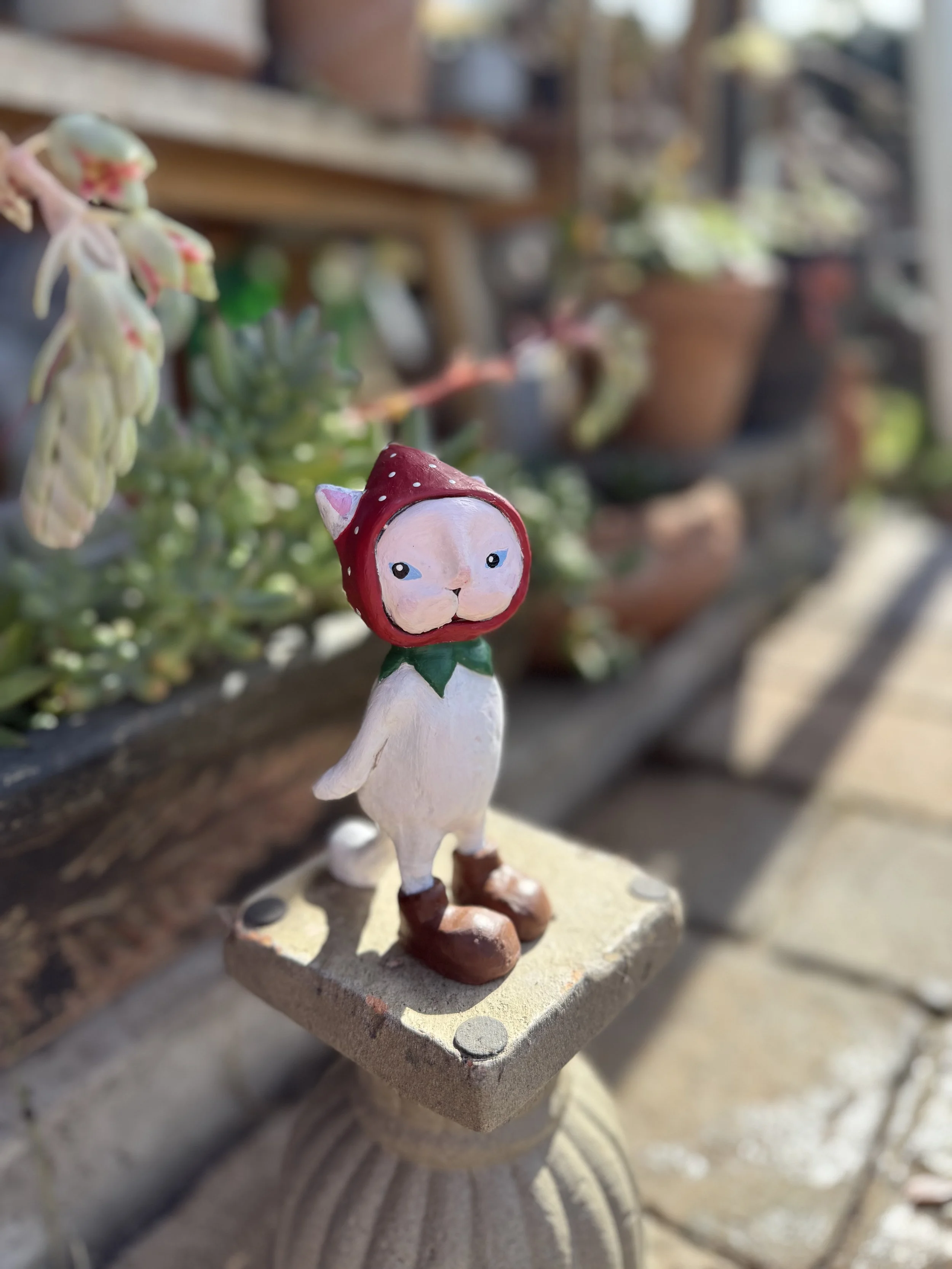 A small, whimsical garden statue of a creature with a white body, brown boots, and a red hat with white dots, standing on a textured pedestal. In the background, potted plants sit on a wooden shelf.