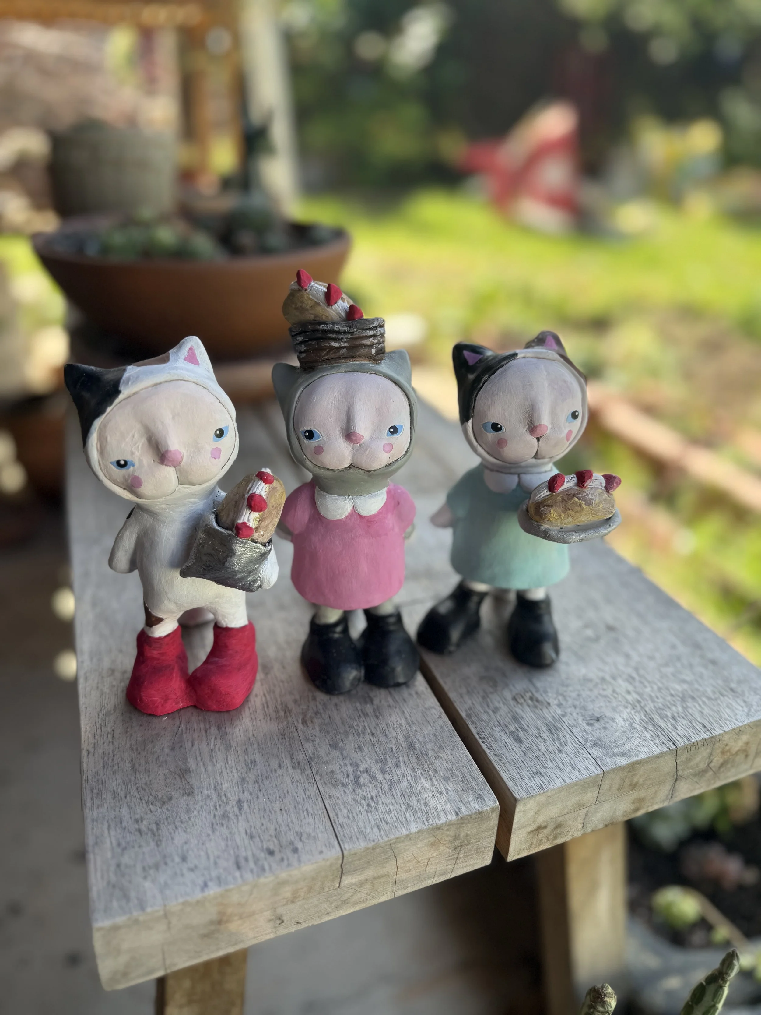 Three small decorated cat figurines with cute faces, each holding or topped with desserts, placed on a weathered wooden table outdoors, with a blurred garden background.