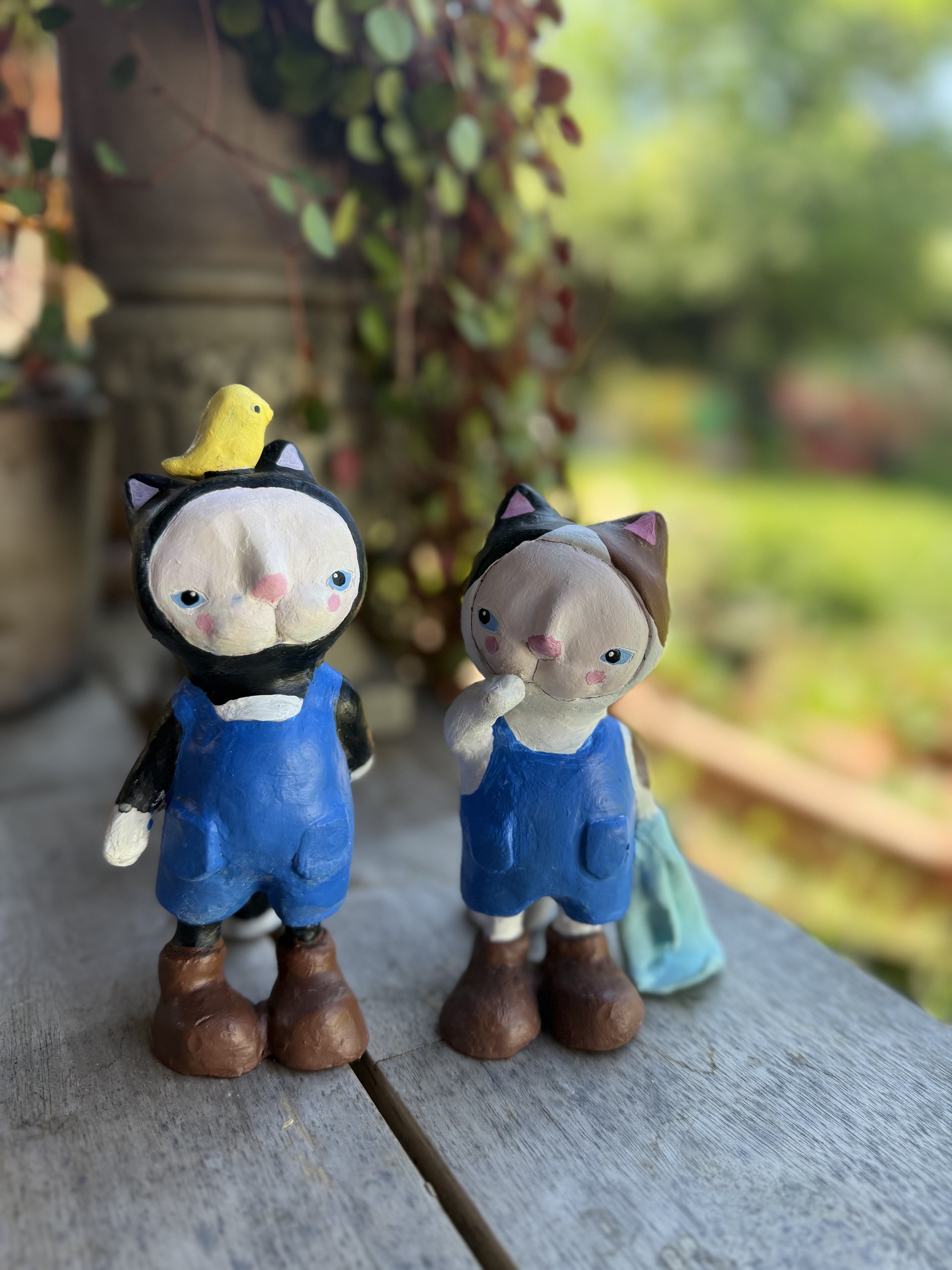 Two ceramic cat figurines dressed in blue overalls standing on a wooden surface outdoors with blurred greenery in the background. A small yellow bird figurine is perched on the head of the left cat.