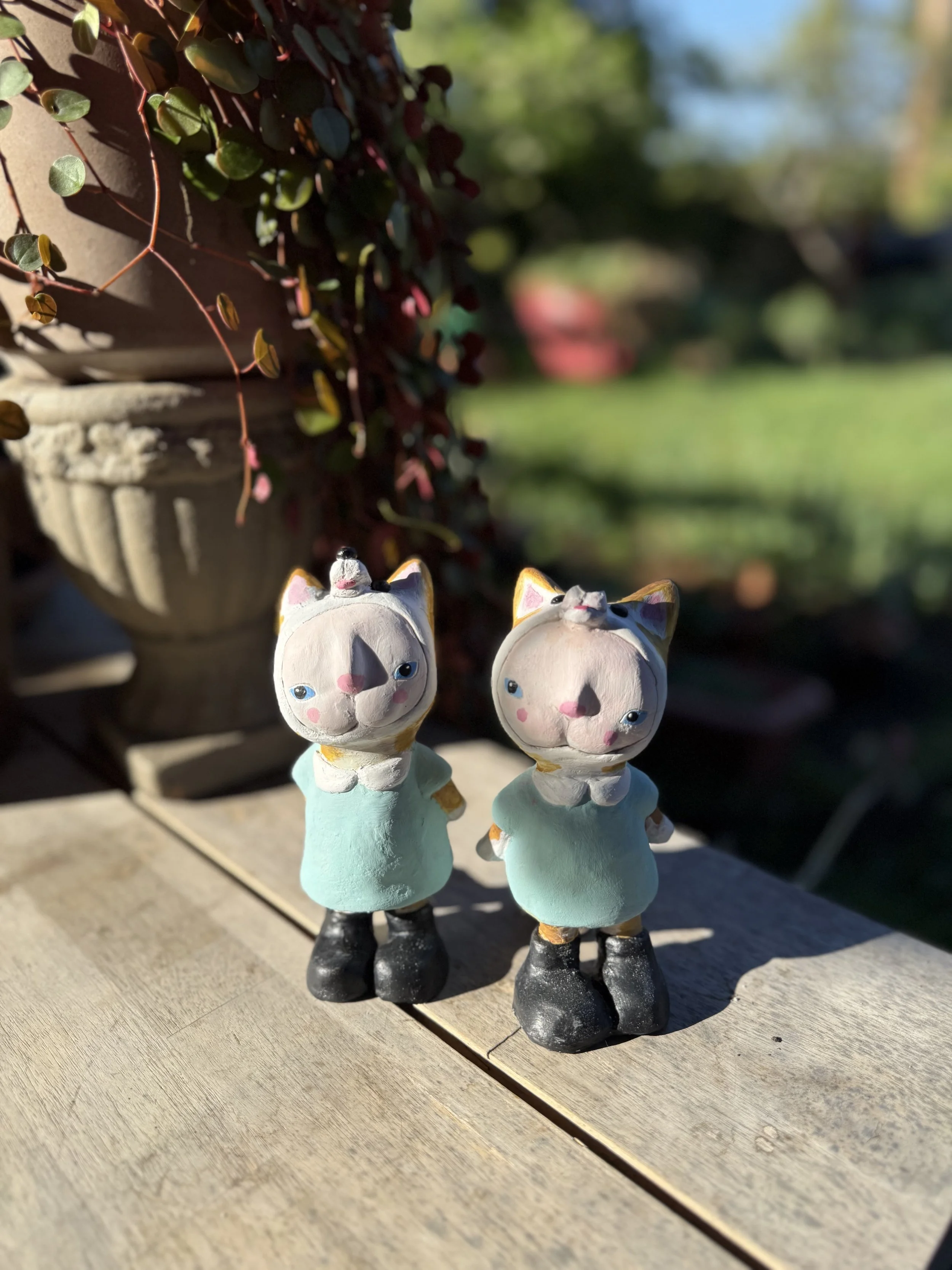 Two ceramic cat figurines standing on a wooden surface outside in bright sunlight, with a flower pot and red and green foliage in the background.