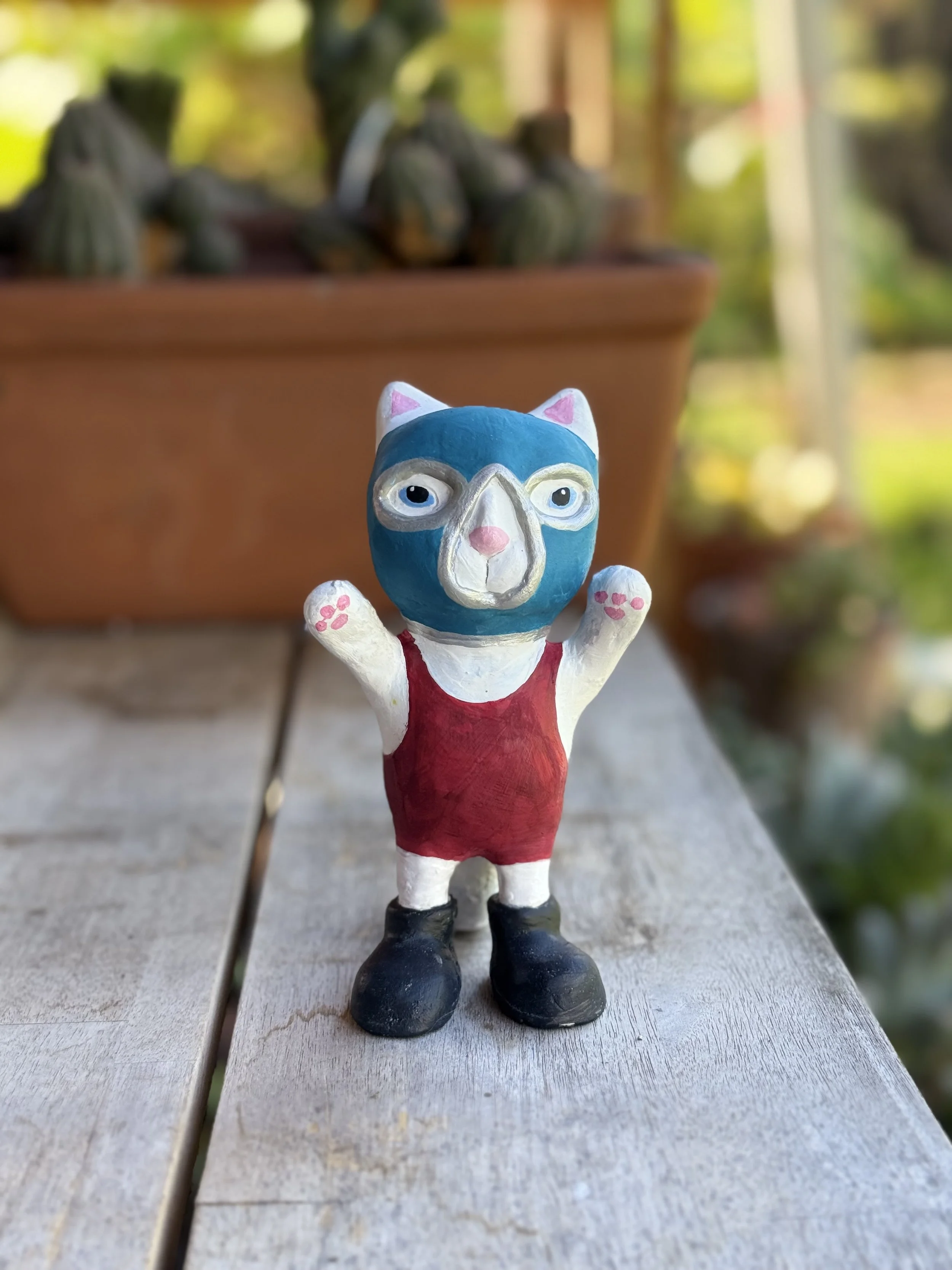 Colorful figurine of an animal with a bear-like head, wearing a red outfit and black shoes, standing on a weathered wooden surface outdoors.