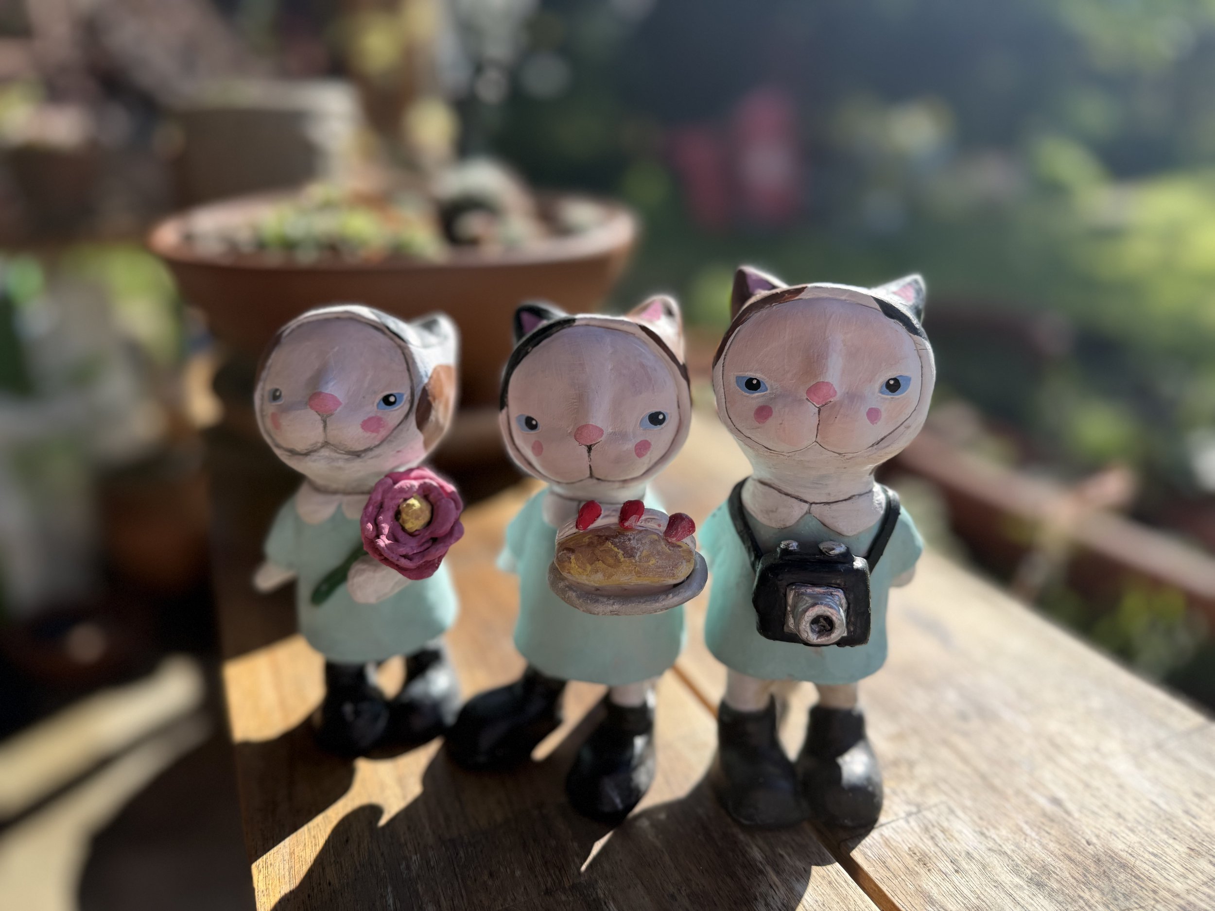 Three decorative cat figurines standing on a wooden surface outdoors, each wearing teal dresses and holding different items: a pink flower, a cake, and a camera.