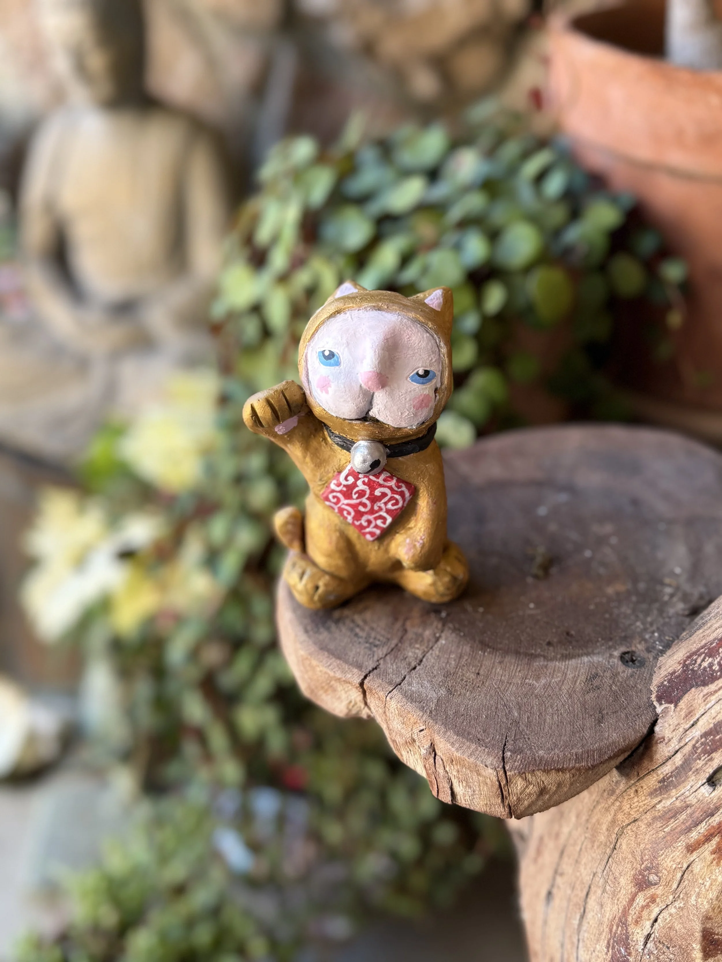 A small figurine of a cat with blue eyes, pink cheeks, and a black collar with a bell and red patterned bandana, standing on a wooden surface with a planter and greenery in the background.