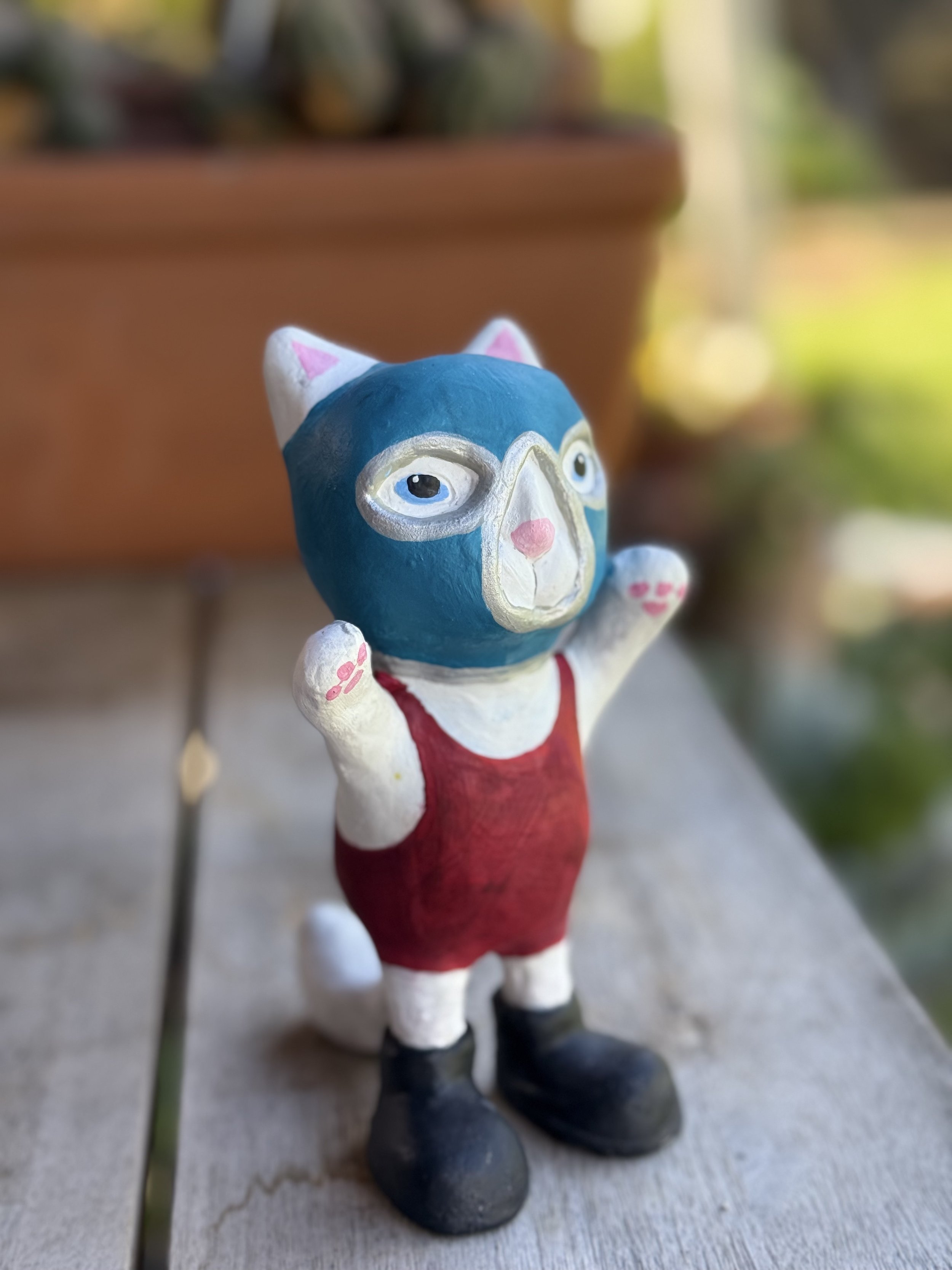 A small, painted figurine of an anthropomorphic cat with glasses, a red dress, and black boots, standing outdoors on a wooden surface with a blurred background of greenery and a large flowerpot.