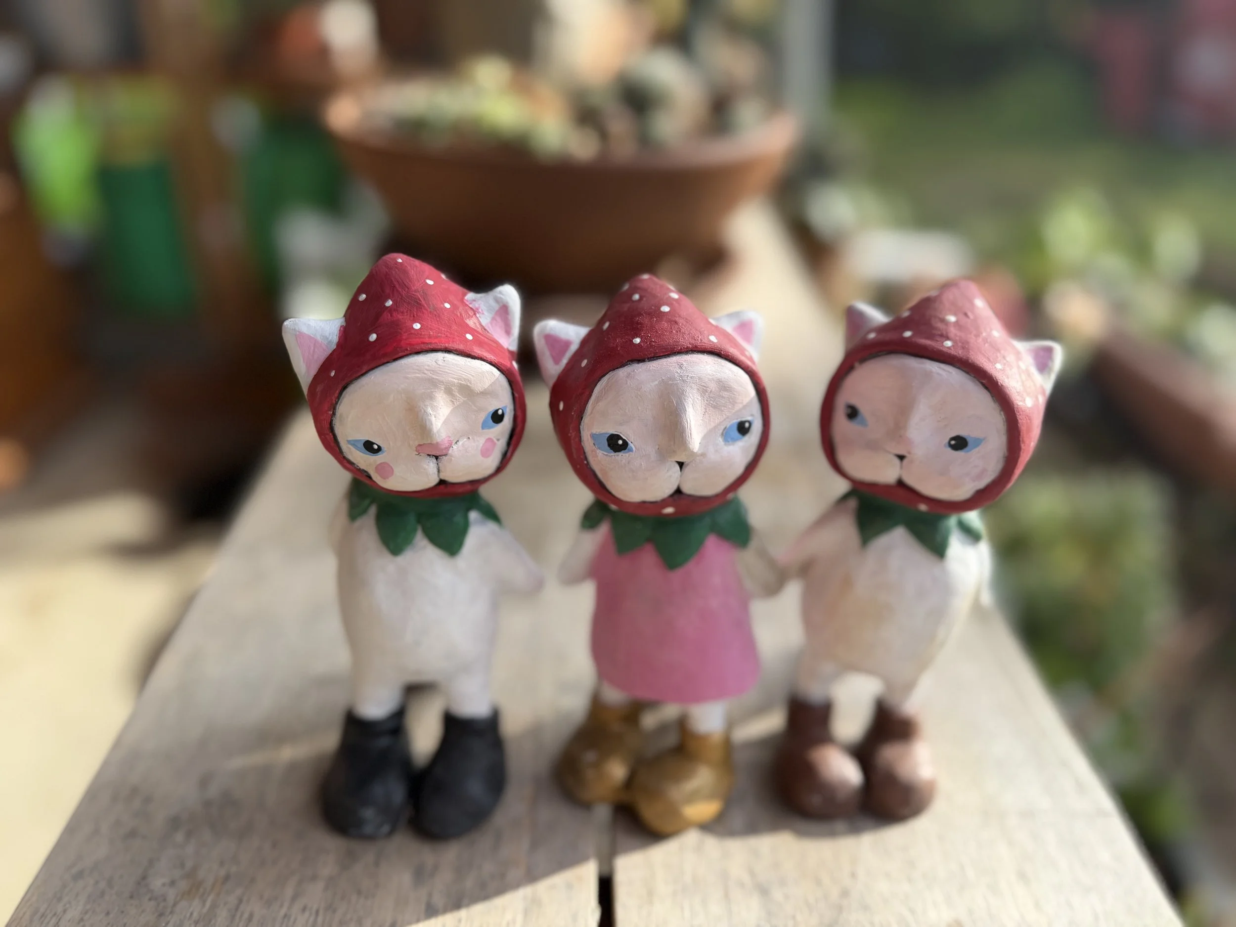 Three small ceramic figurines of cats in strawberries costumes, each with a green collar and elf-like shoes, standing on a wooden surface with blurred background.