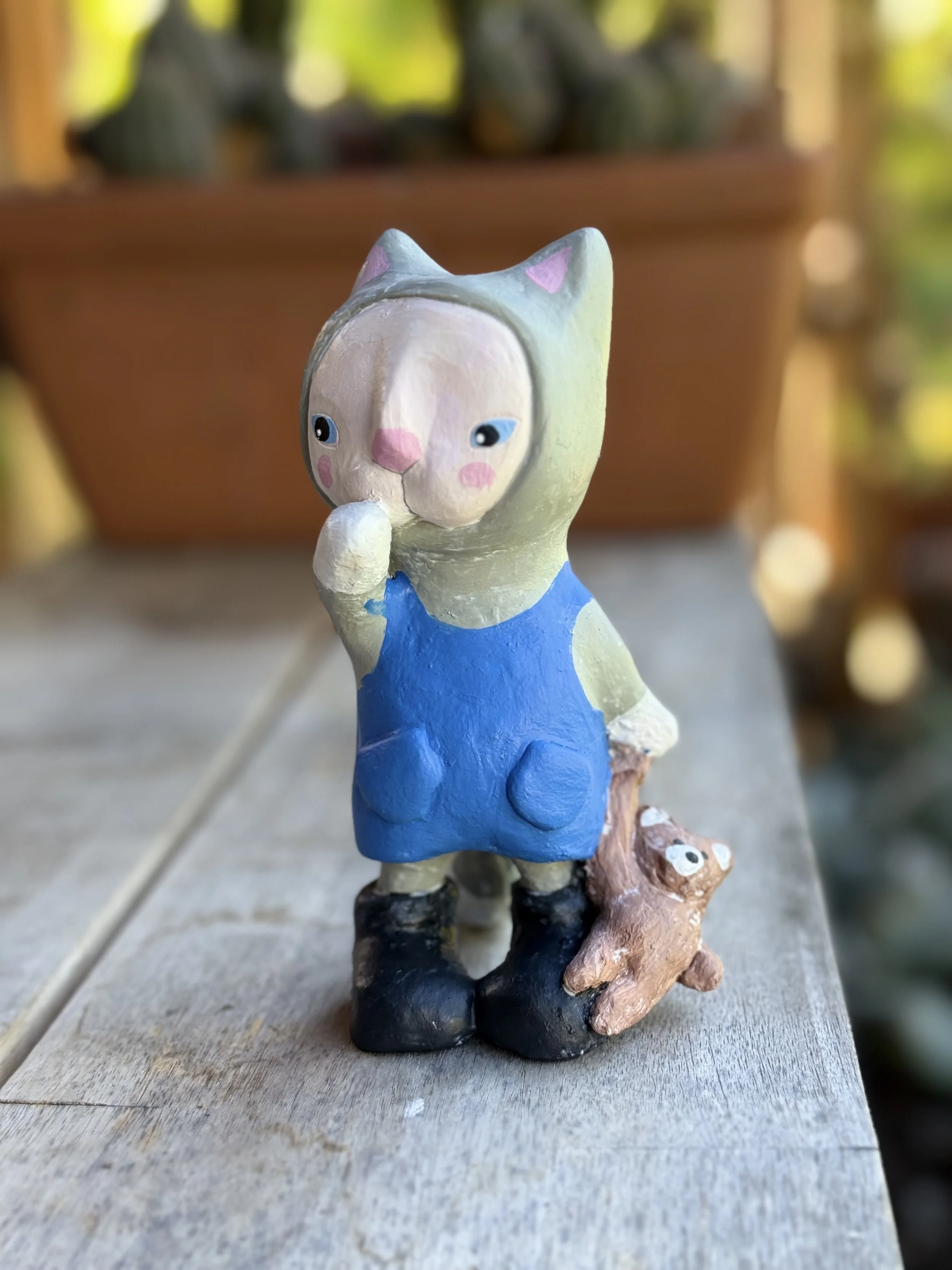 A small figurine of a girl with a cat costume on her head, wearing a blue dress, standing on a wooden surface with a small brown animal at her feet, outdoors with blurred green plants in the background.