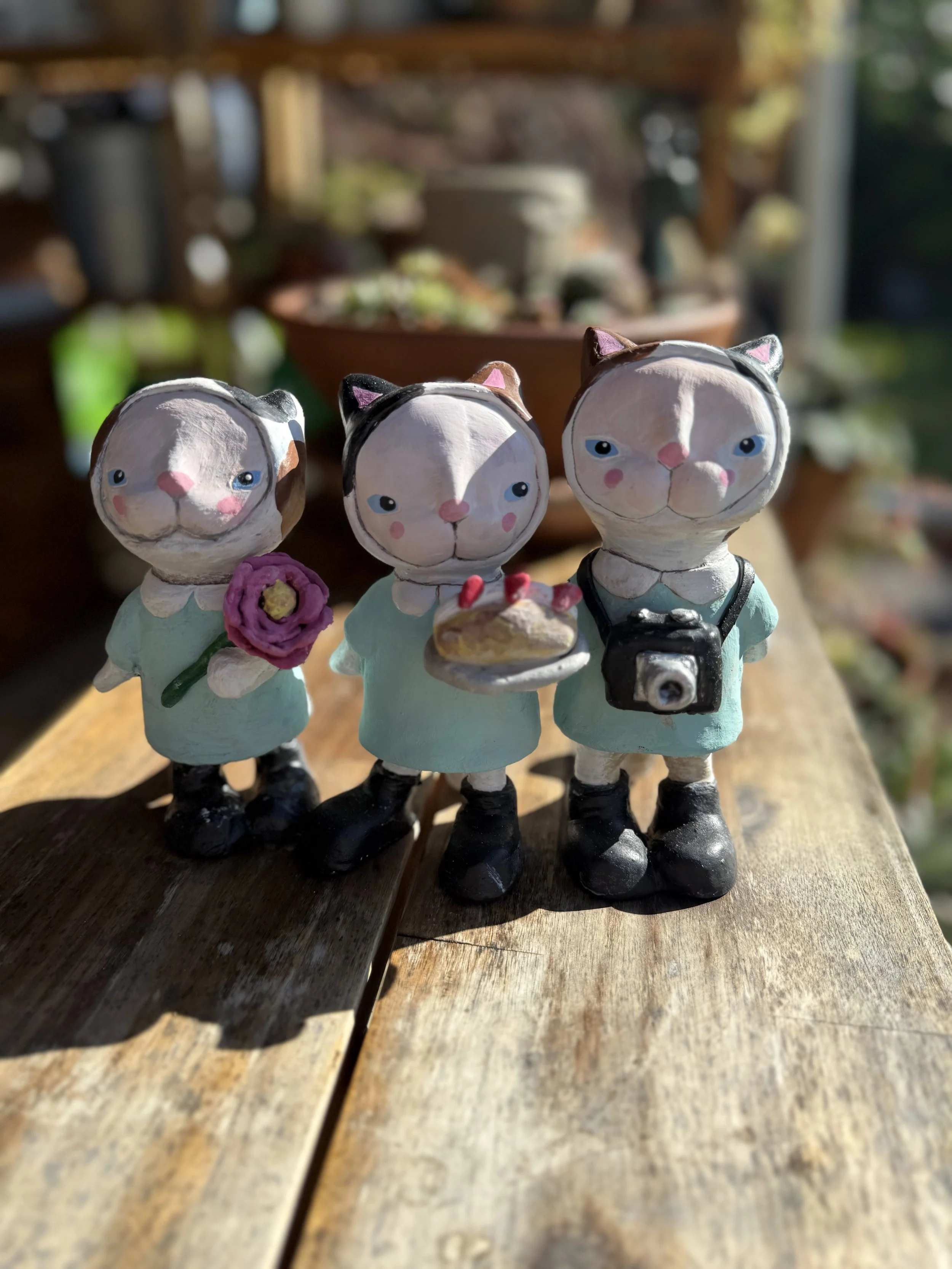 Three cat figurines dressed as girls, each with different accessories, standing on a wooden surface outdoors in sunlight.