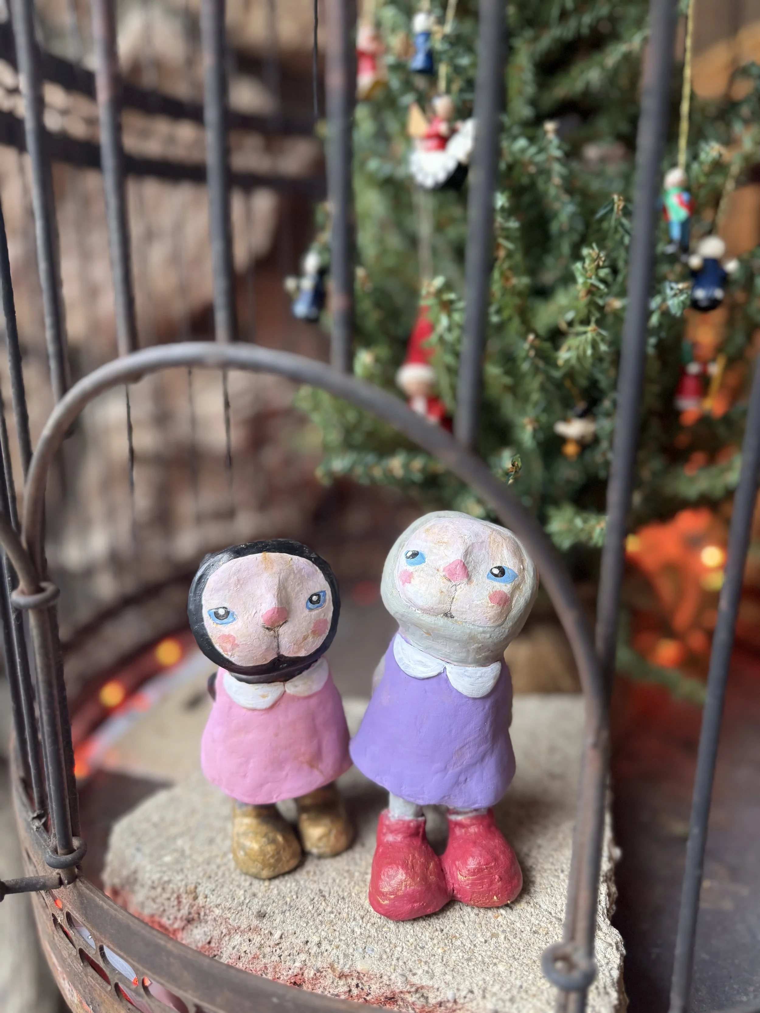Two hand-painted ceramic figurines of girls with rosy cheeks, standing inside a metal cage, with a decorated Christmas tree in the background.
