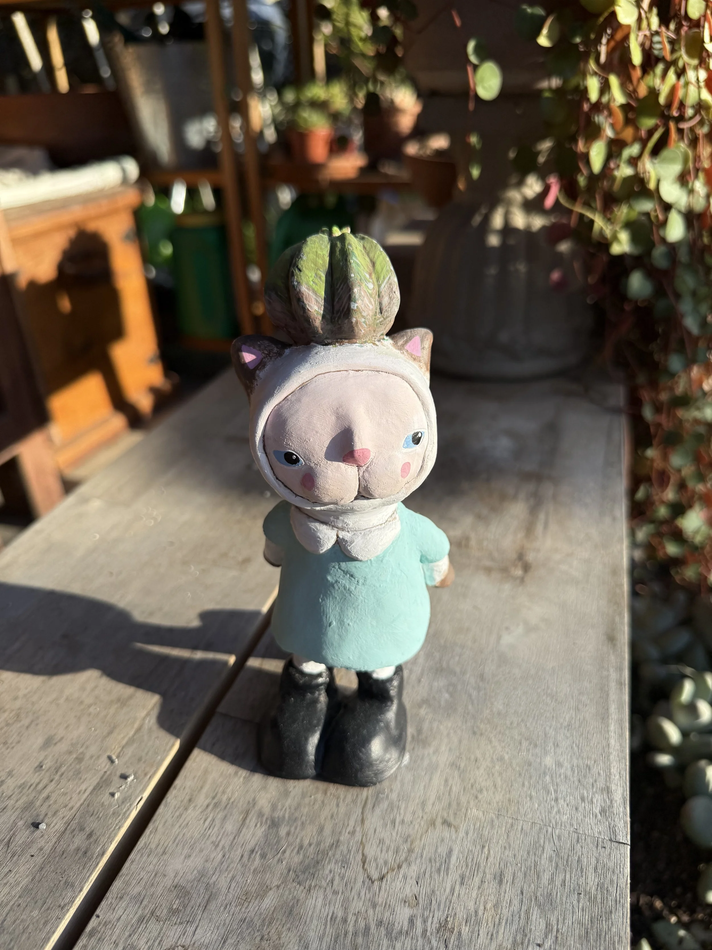 A small, whimsical figurine of a girl with a cat-shaped hat, standing on a wooden surface outdoors. The figurine has a large head with blue eyes, pink cheeks, and black boots. The background features blurred plants and garden furniture.
