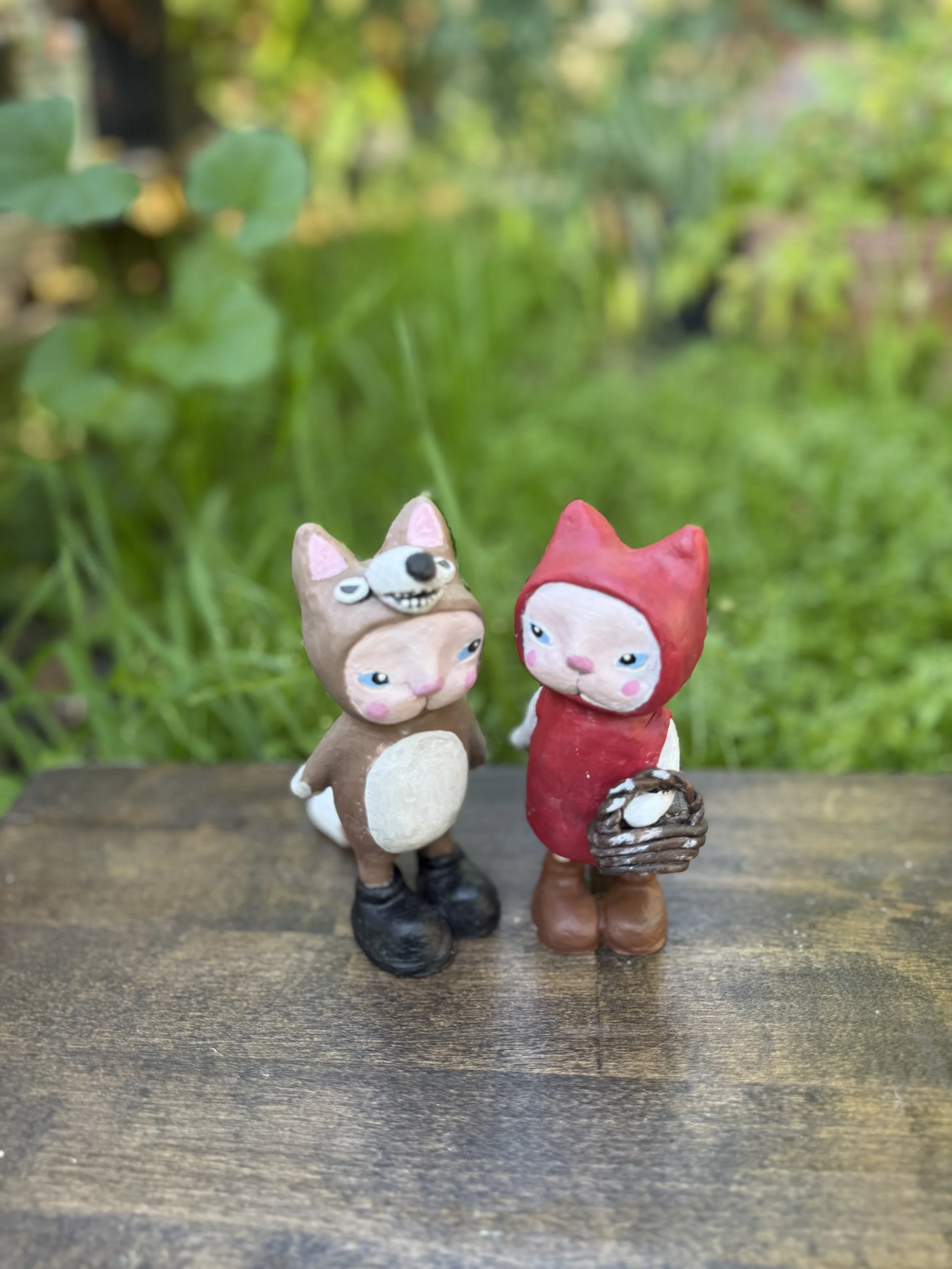 Two handmade figurines of animals, one dressed as a dog and the other as a cat, standing on a wooden surface outdoors with greenery in the background.