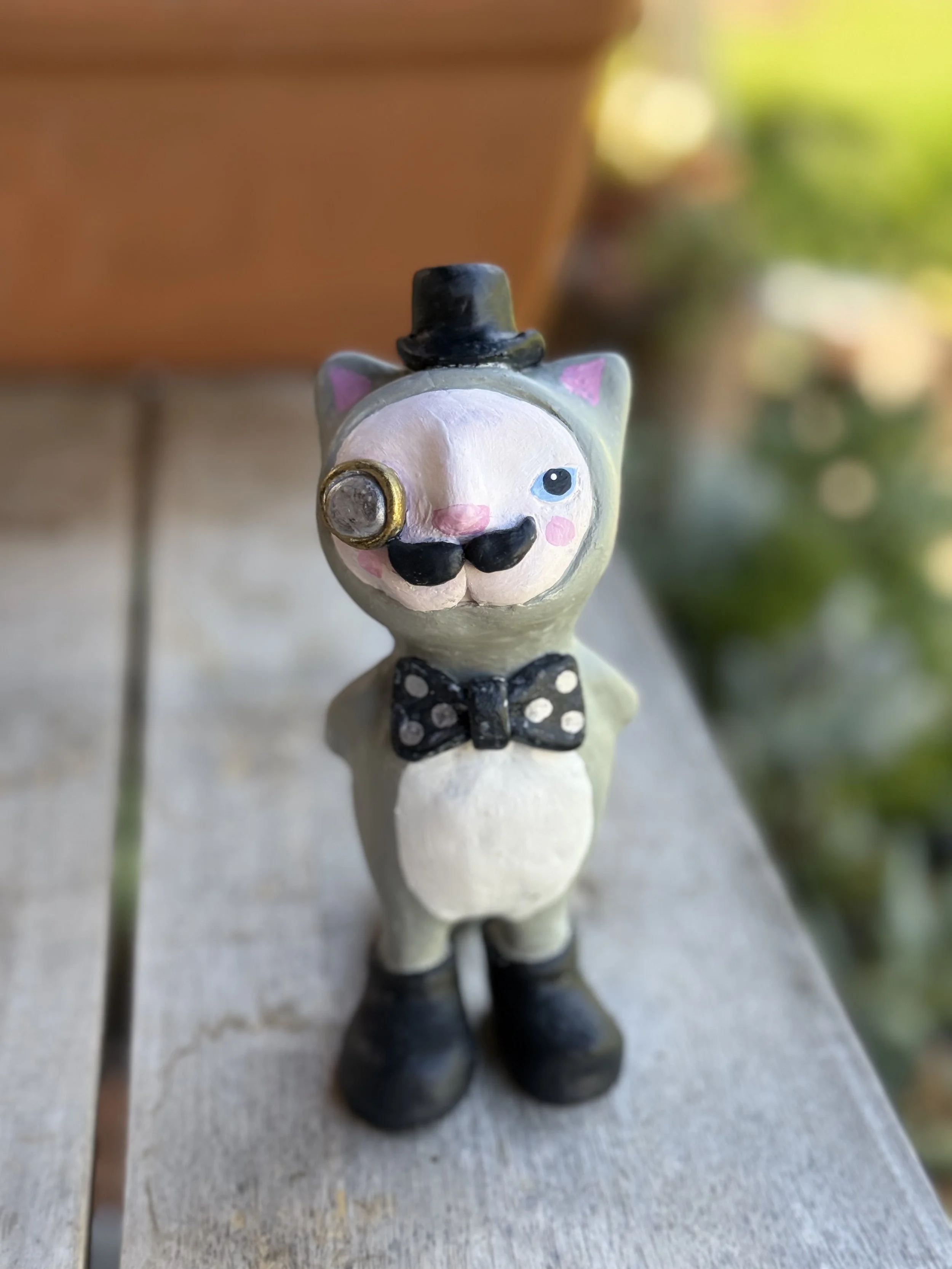 Whimsical toy figure of a cat dressed as a gentleman, wearing a top hat, bow tie, and boots, with a monocle over one eye, on a wooden surface outdoors.