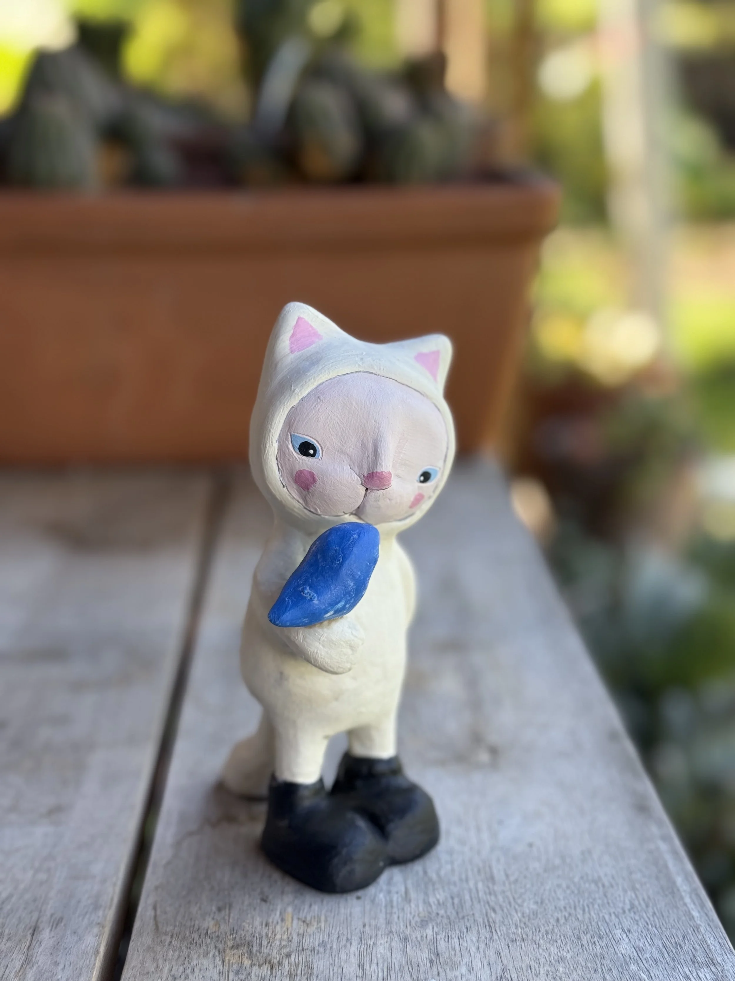 A small figurine of a sheep with a human-like face wearing a white hood with pink ears, black boots, and holding a blue object, standing on a wooden surface with a blurred outdoor background.