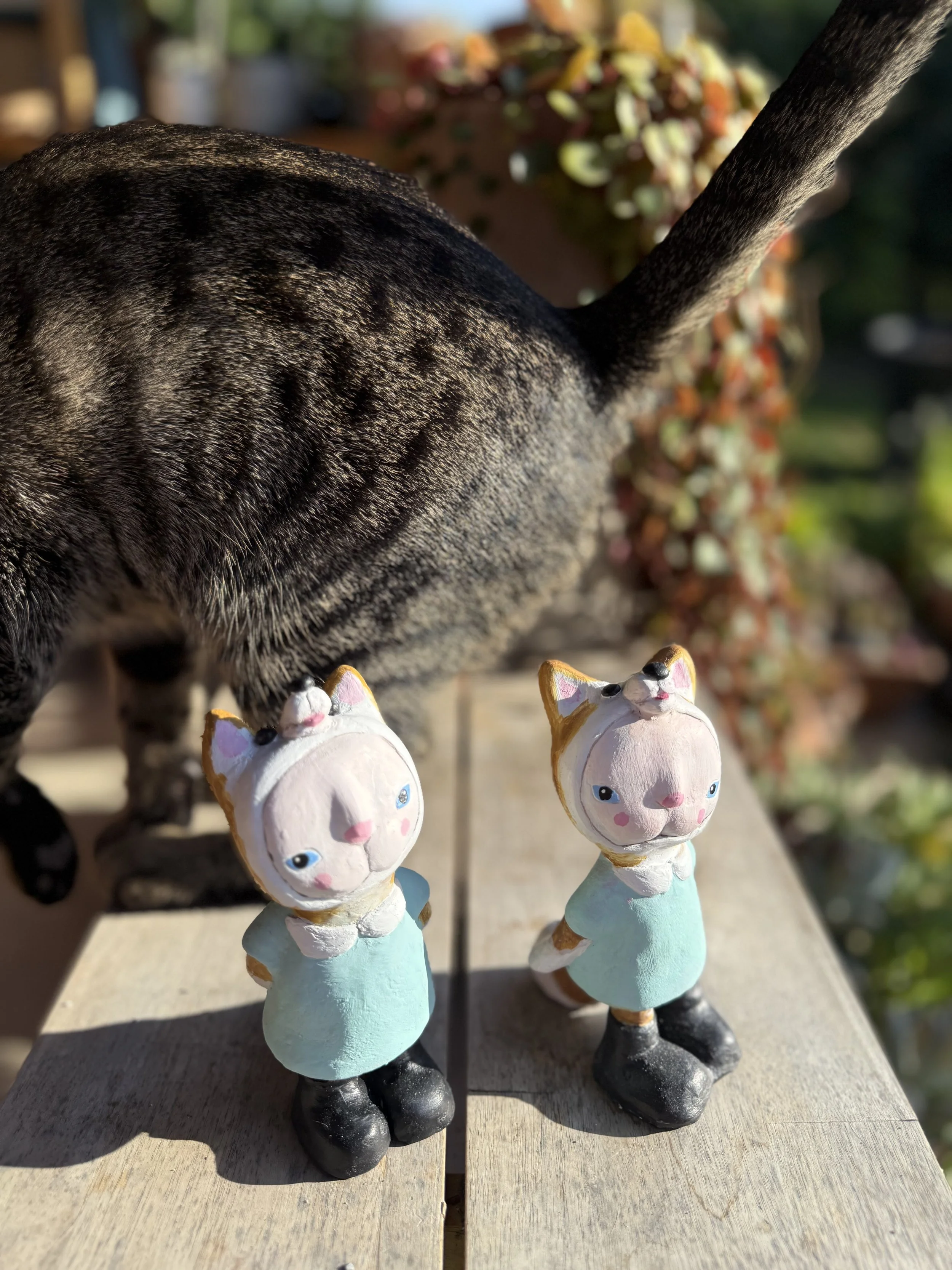 Two small figurines of cats dressed in light blue dresses and black boots, with a cat's tail visible in the background. The figurines are positioned on a wooden surface outdoors, with a blurred background of greenery and flowers.