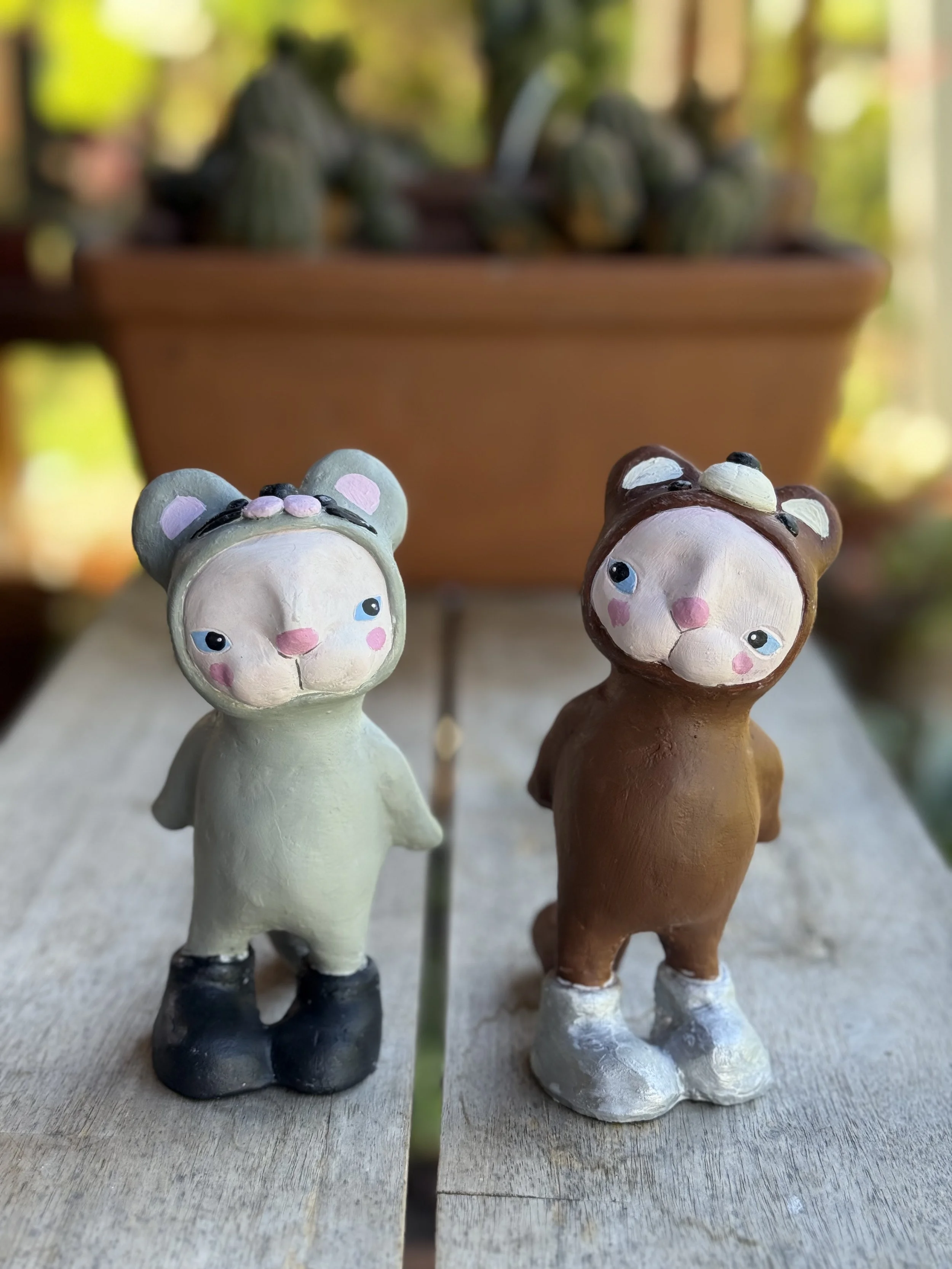 Two animal figurines, one resembling a mouse and the other a bear, standing on a wooden surface with a blurred plant and flowerpot in the background.