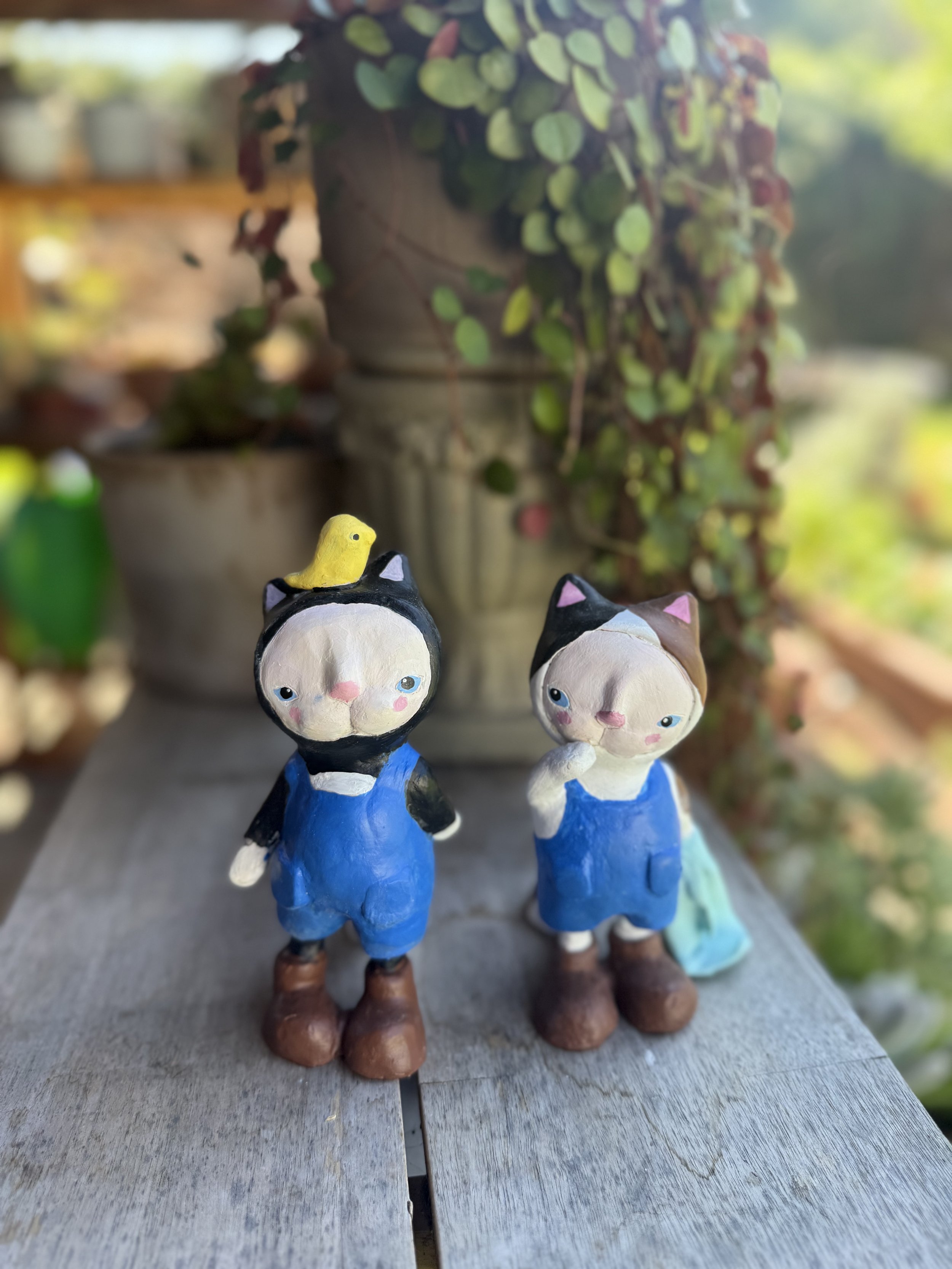 Two ceramic figurines of cats dressed in blue overalls, with one cat having a yellow chick on its head, placed on a weathered outdoor wooden surface with potted plants and greenery in the background.