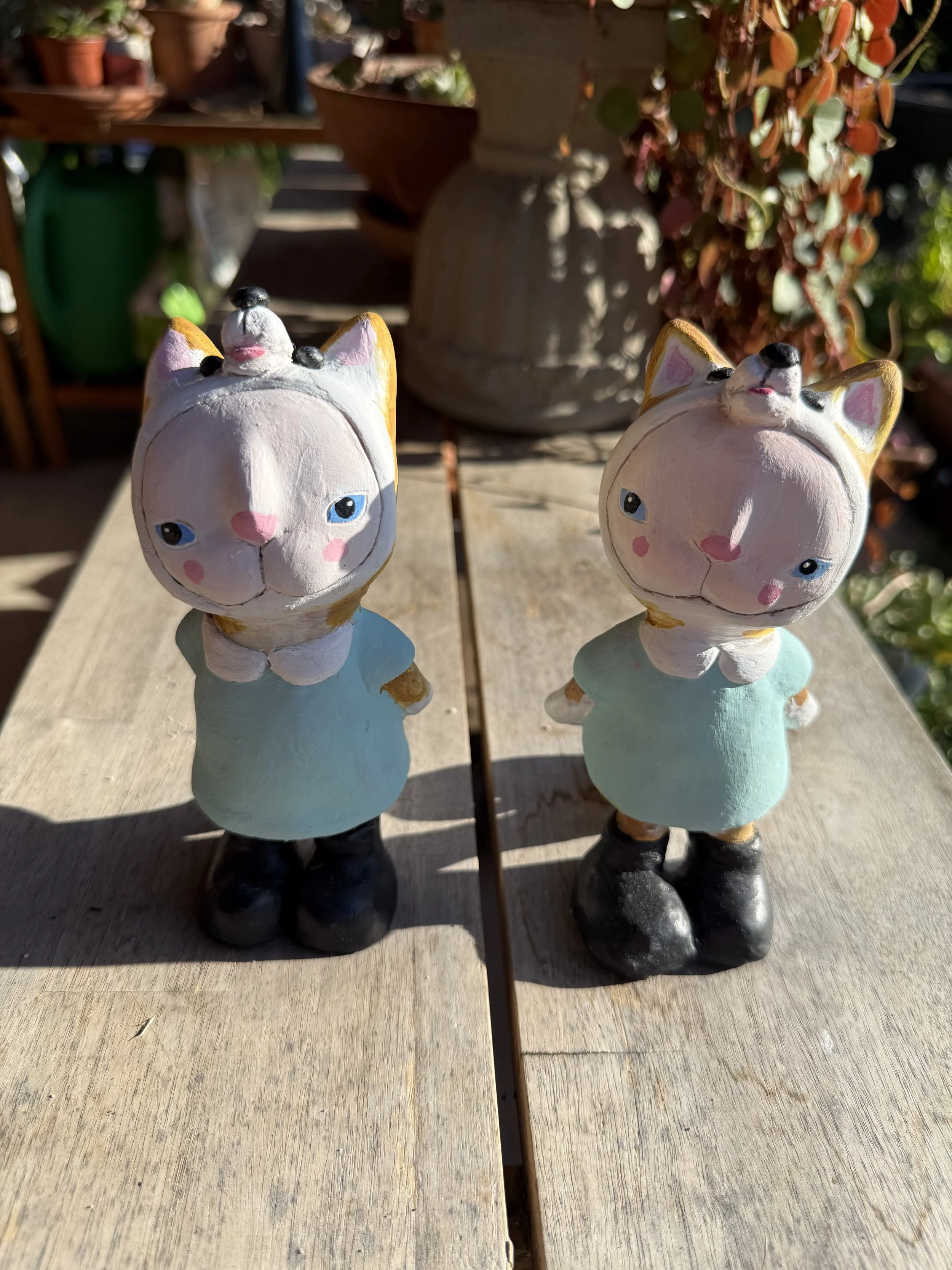 Two ceramic cat figurines with pink cheeks, one with a small bird on its head, standing outdoors on a wooden surface with leafy plants and a large planter in the background.