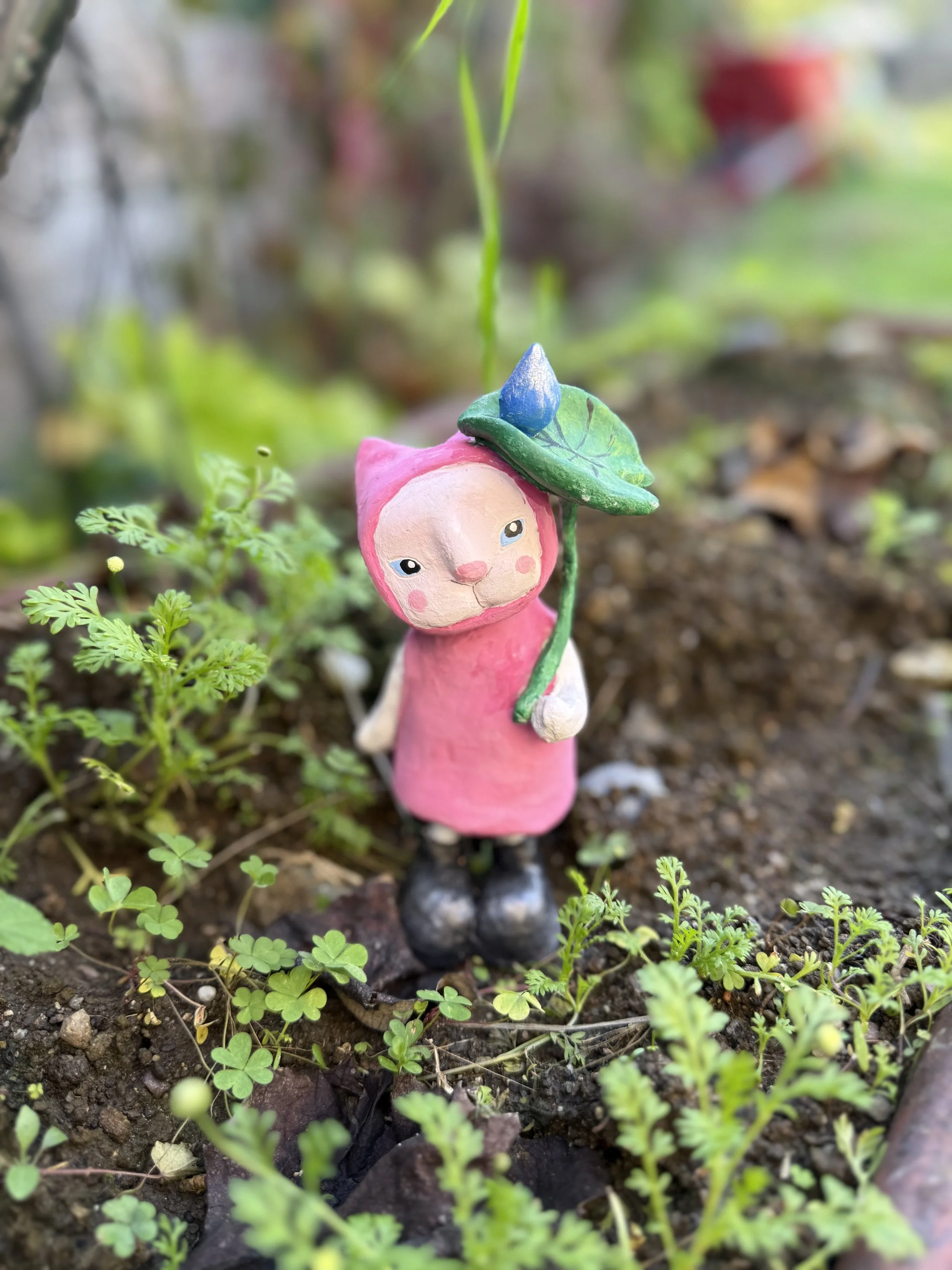 A small, whimsical figurine of a pink creature with a cat-like face, standing in a garden among green plants. The figurine is wearing a pink hooded cloak and black shoes, and it holds a green leaf with a blue bud on top.