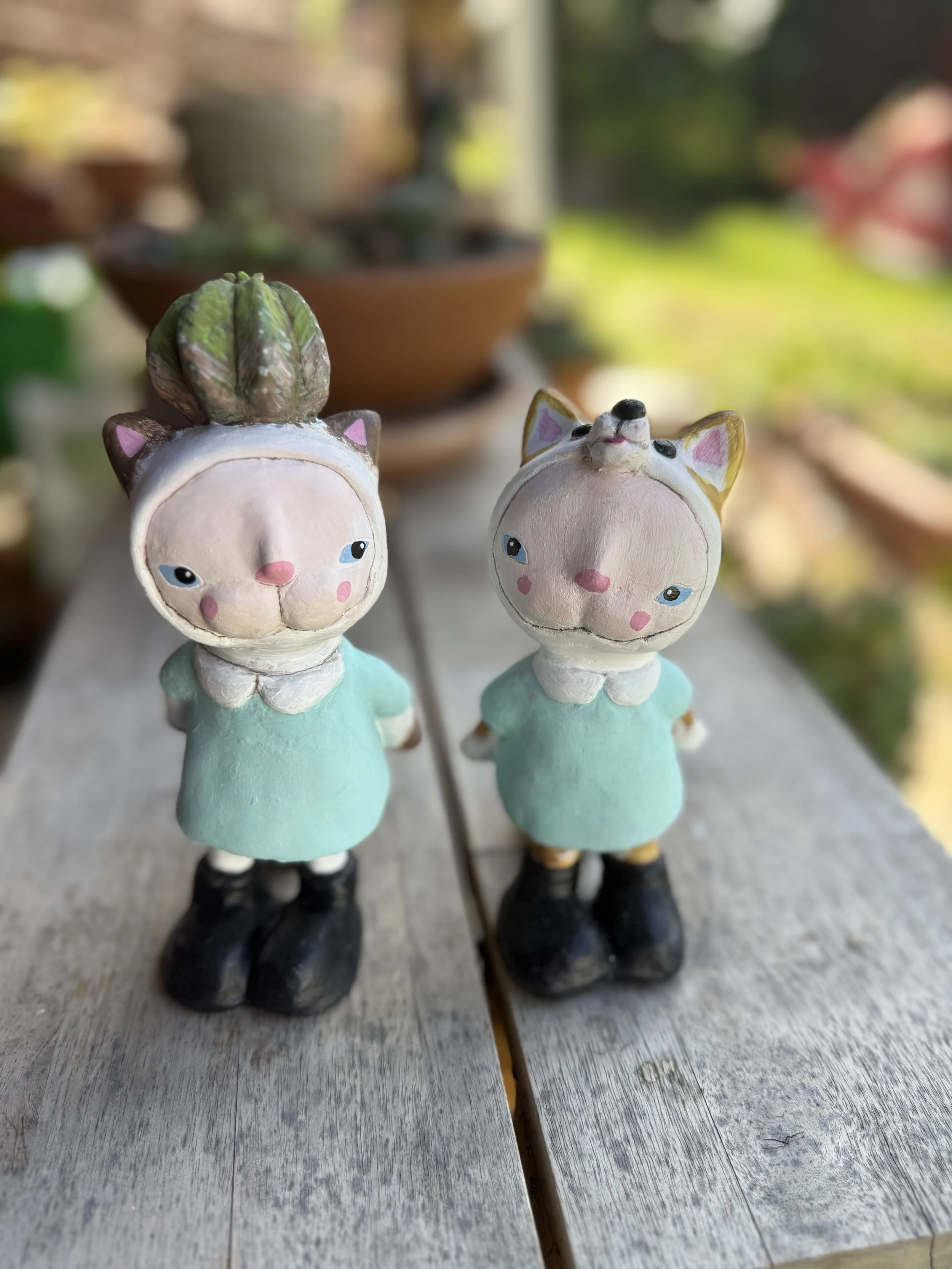Two decorative animal figurines with human features standing on a wooden surface outdoors, one resembling a cat and the other a dog, both wearing light blue dresses and black shoes.