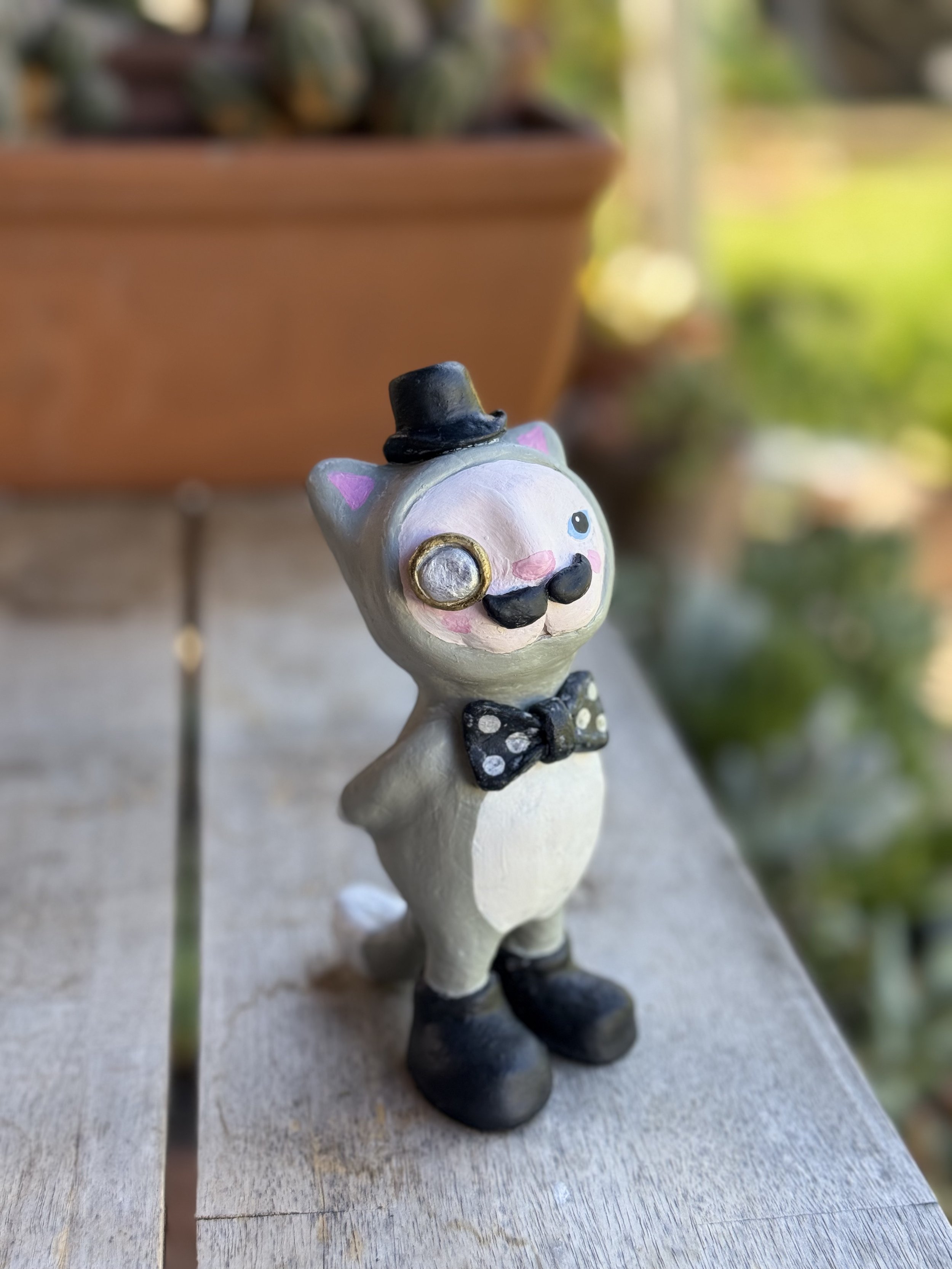 Decorative figurine of a cat dressed as a gentleman, wearing a black top hat, monocle, bow tie, and black boots, standing on a wooden surface outdoors.