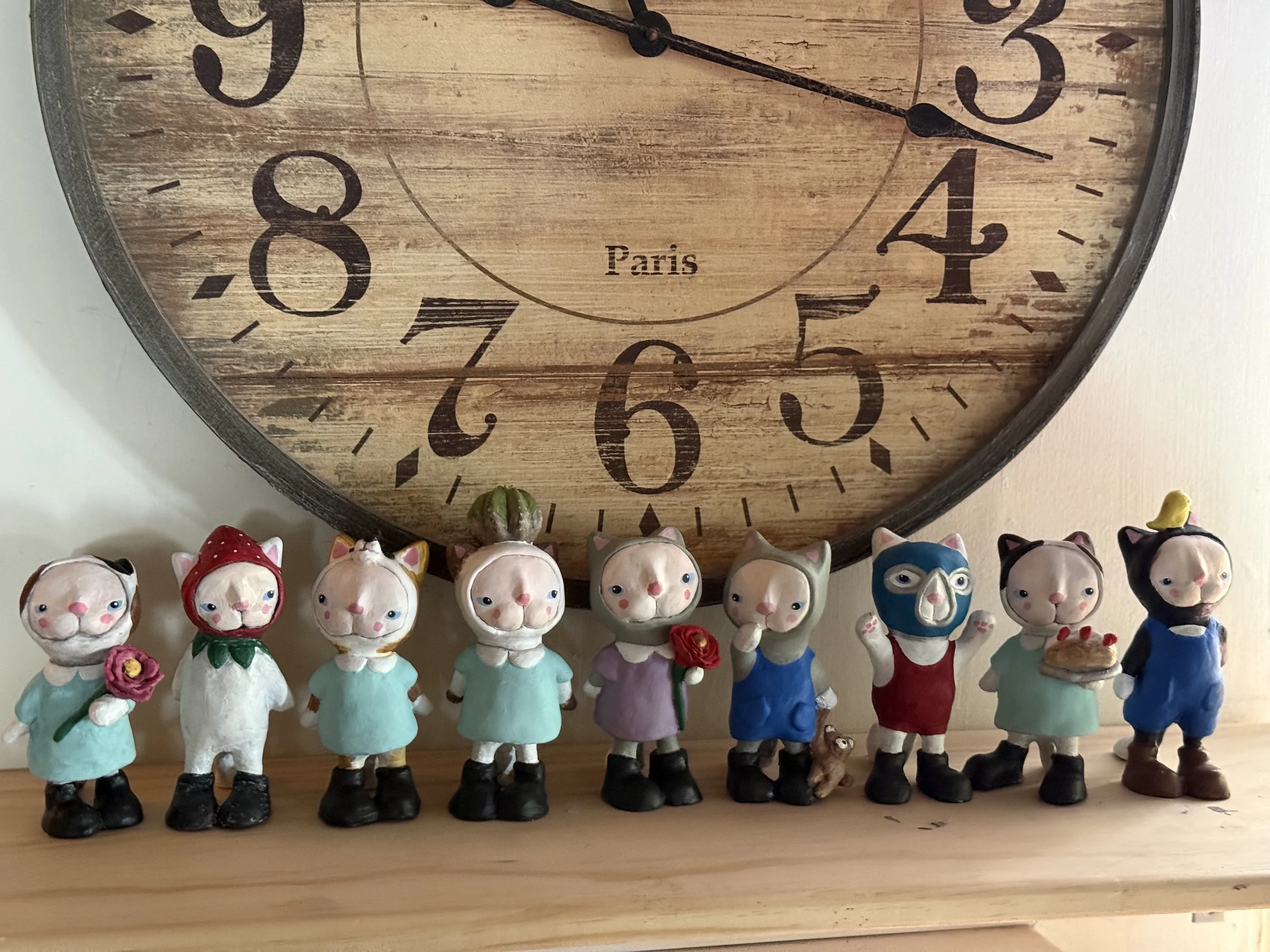 Set of nine small pig figurines dressed in various colorful outfits, standing in a row on a wooden surface in front of a large round wall clock with Roman numerals and the word 'Paris' on the face.