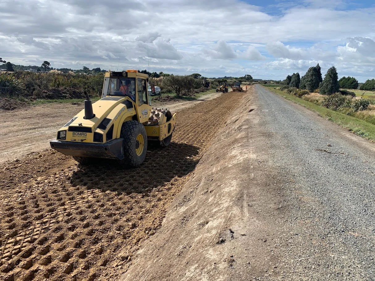Construction workers leveling the dirt roadside along a gravel road with a yellow road roller under a partly cloudy sky.