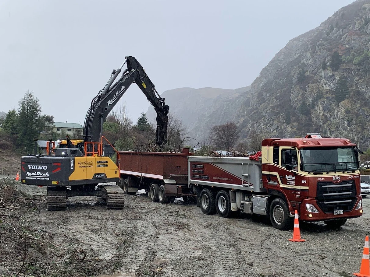 Construction vehicles, including a Volvo excavator and a red dump truck, working together on a muddy outdoor site with mountains in the background, and orange traffic cones marking the area.