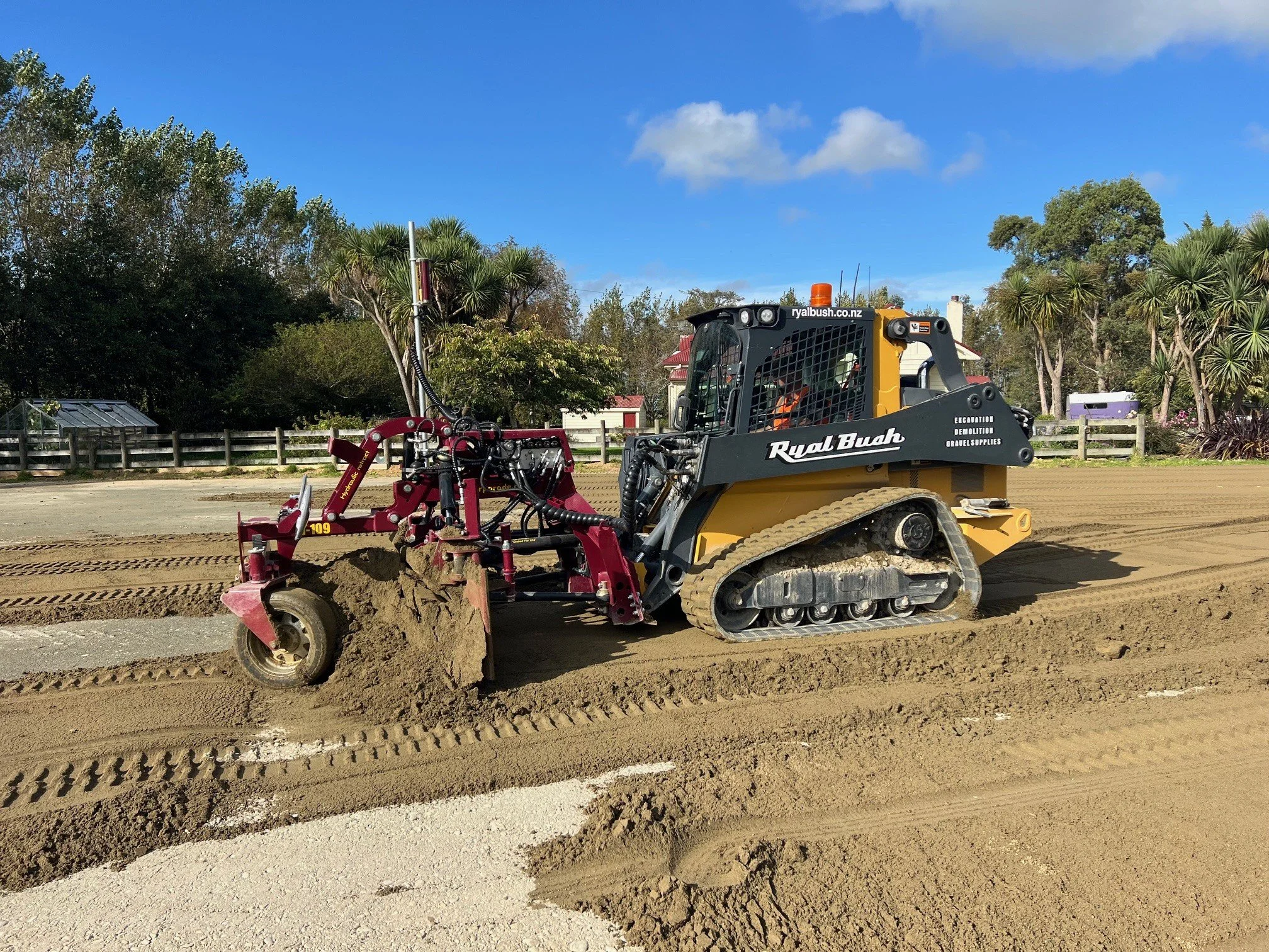 A small tracked construction vehicle, painted yellow and black, is leveling dirt with a red land roller attachment on a construction site. The scene has blue skies and trees in the background.
