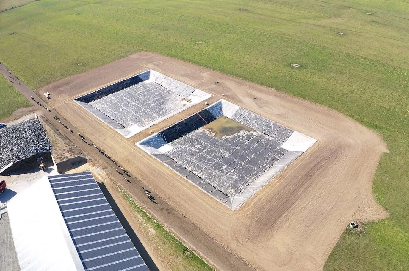Aerial view of two large rectangular fish farming tanks with black liners, surrounded by dirt paths and green fields.